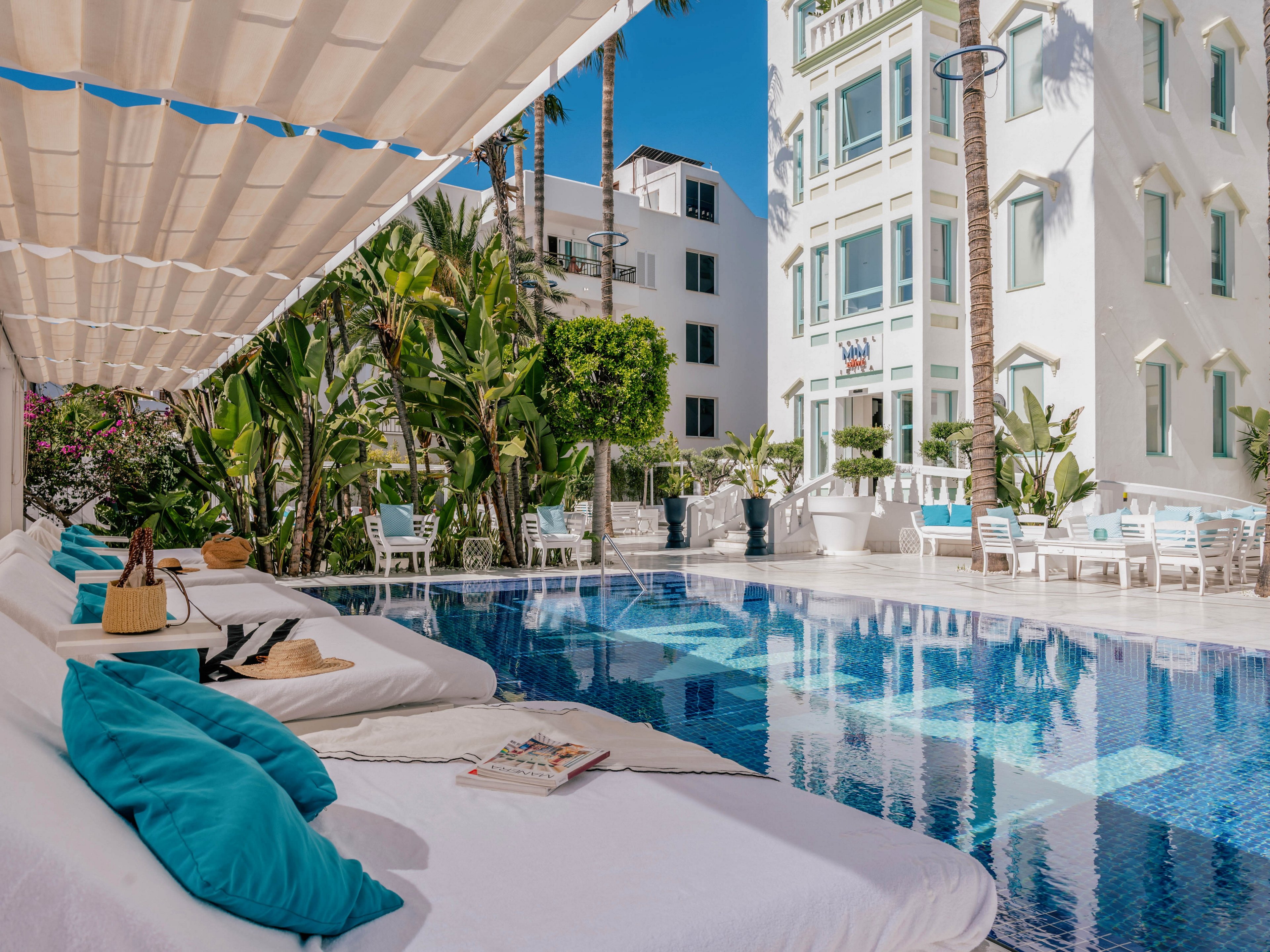 A luxurious hotel pool area features white cushioned loungers with bright turquoise pillows under a shaded canopy. The clear blue swimming pool reflects the surrounding white buildings and tall palm trees. Guests relax at white tables and chairs in the background, creating a serene resort atmosphere. Tropical plants and modern architecture enhance the feeling of an upscale vacation retreat.