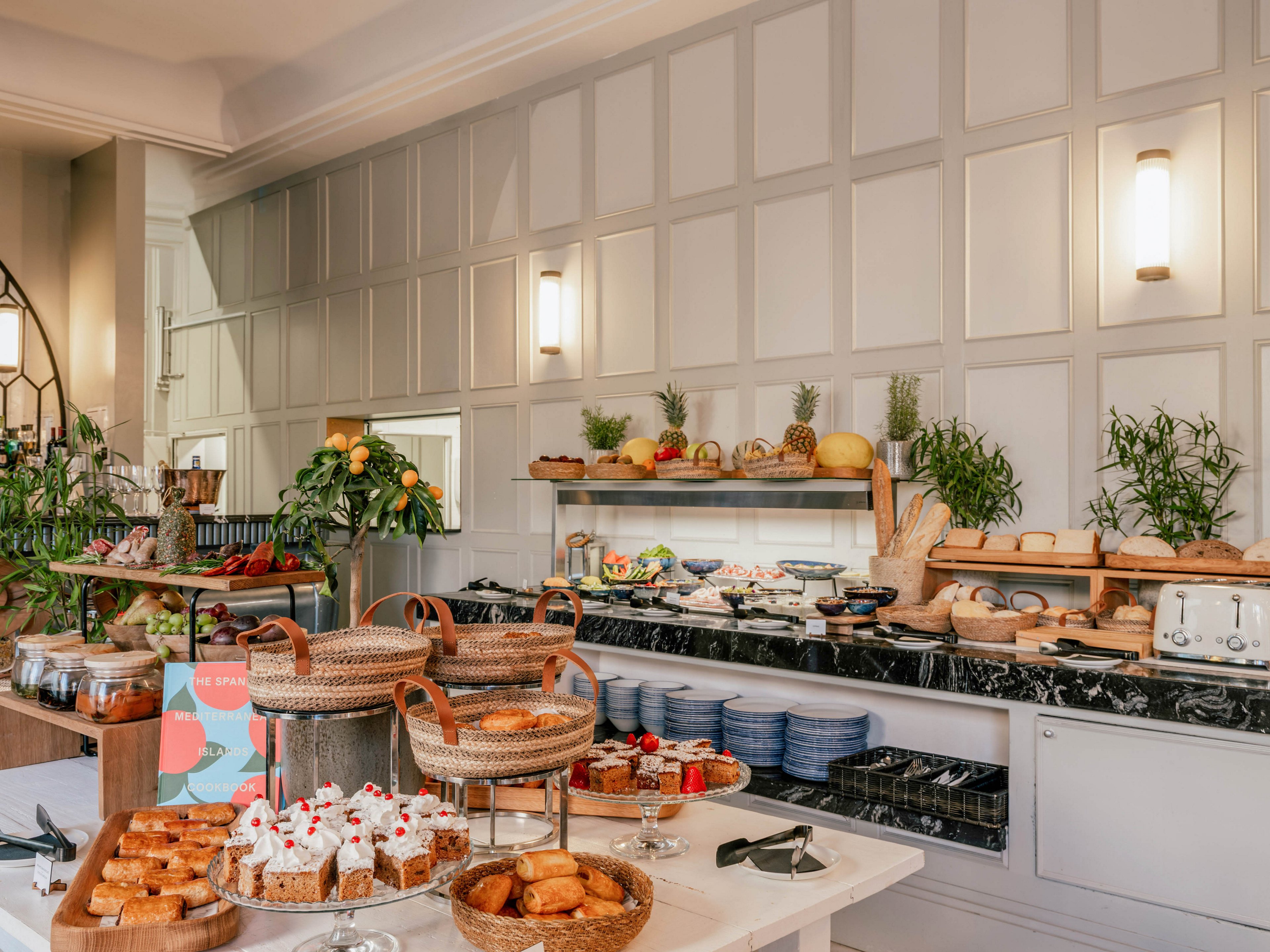 A stylish breakfast buffet is arranged on a long counter in a bright dining room. Various pastries, cakes, and breads are displayed in wicker baskets and on wooden boards. Fresh fruits, salads, and hot dishes are set out in trays and chafing dishes along the back counter. The space features light paneled walls, modern wall sconces, and decorative greenery for a welcoming atmosphere.