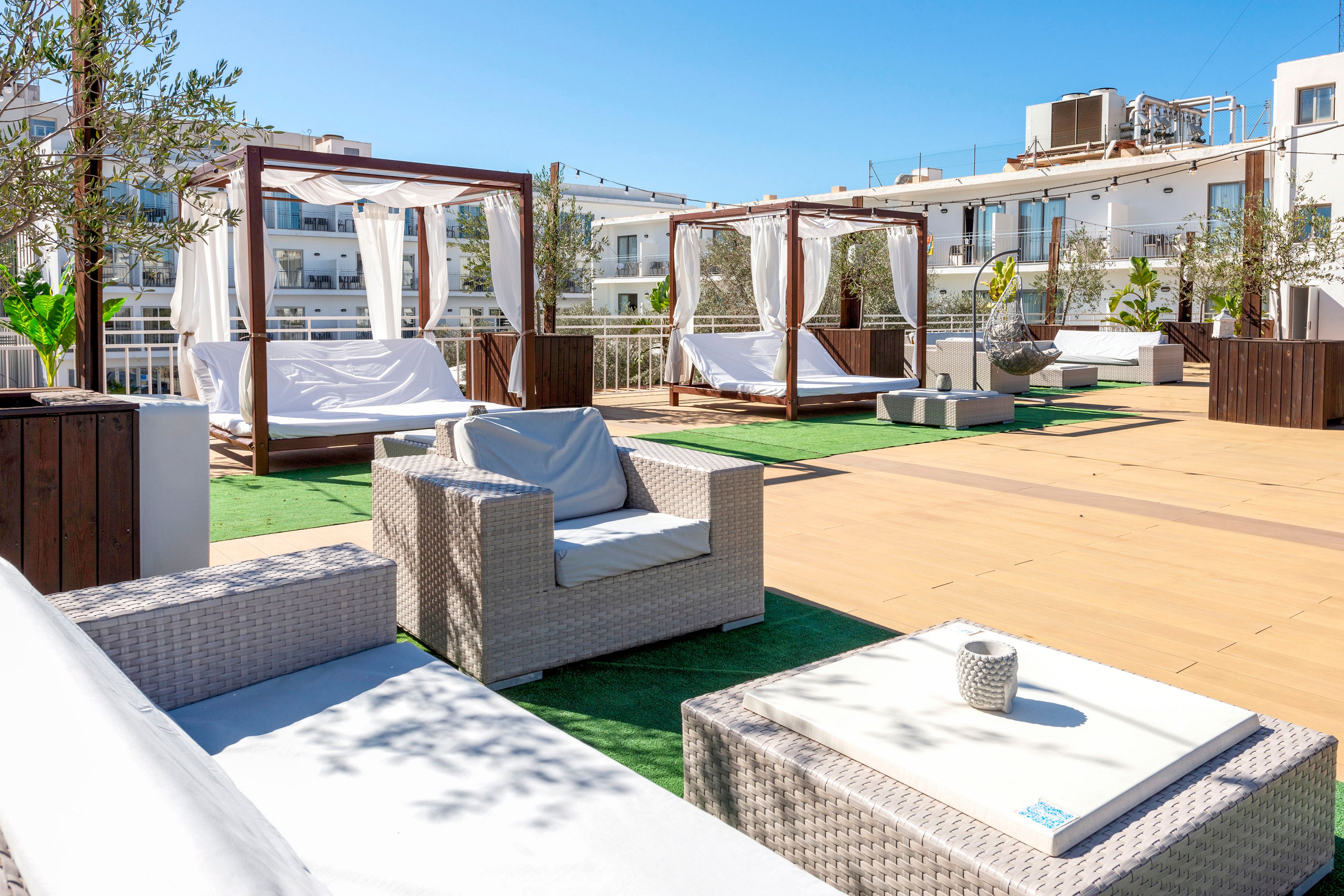 A modern rooftop terrace features wicker lounge furniture and white cushioned seating under a clear blue sky. Several cabana-style daybeds with flowing white curtains are arranged on a spacious deck. Green artificial grass sections contrast with the warm wooden flooring and surrounding white buildings. Potted trees and plants add a touch of nature to the contemporary outdoor relaxation area.