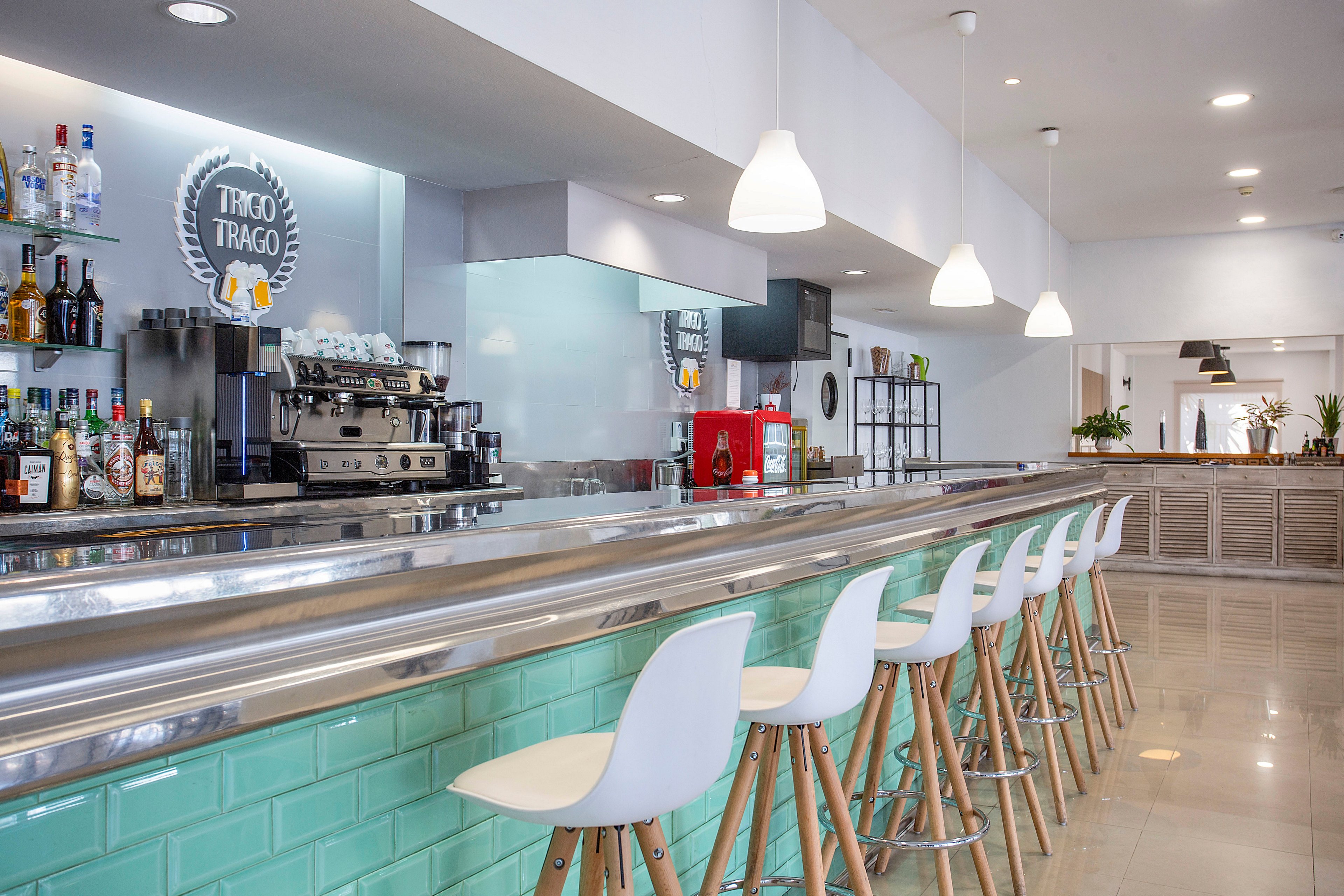 Contemporary bar interior featuring a long metal counter lined with white stools on wooden legs. The front of the bar is finished with glossy turquoise tiles, adding a fresh accent to the neutral space. Shelves behind the counter display assorted bottles and a professional espresso machine beneath a circular sign reading "TRAGO TRAGO". Pendant lights and polished flooring create a bright, clean atmosphere with plants visible in the background.