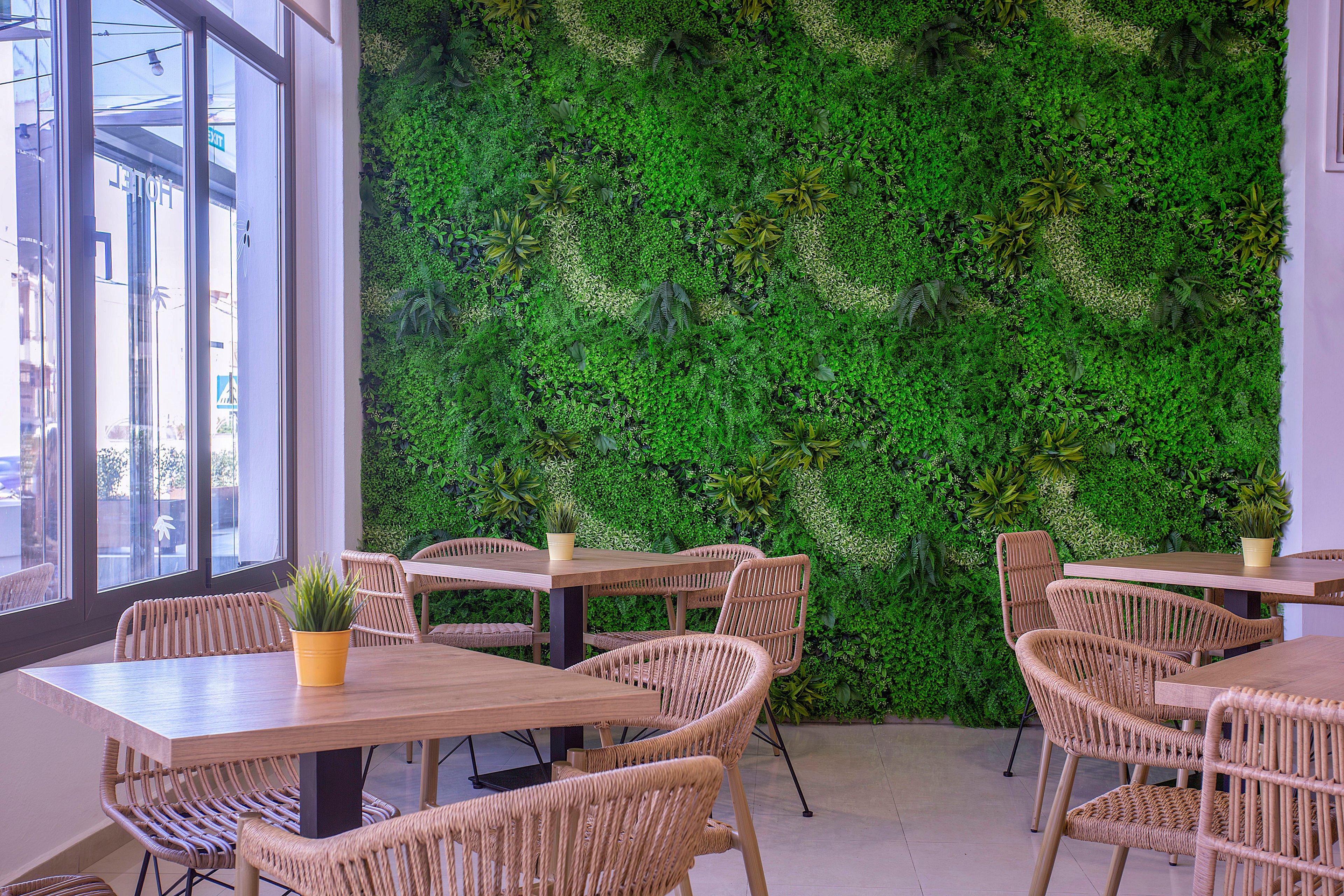 Contemporary café seating area features wooden tables and woven rattan chairs. A vibrant vertical green wall of plants dominates the background, creating a fresh, natural atmosphere. Small potted plants sit on the tables, reinforcing the botanical theme. Large windows on the side bring in daylight and offer a view of the street outside.