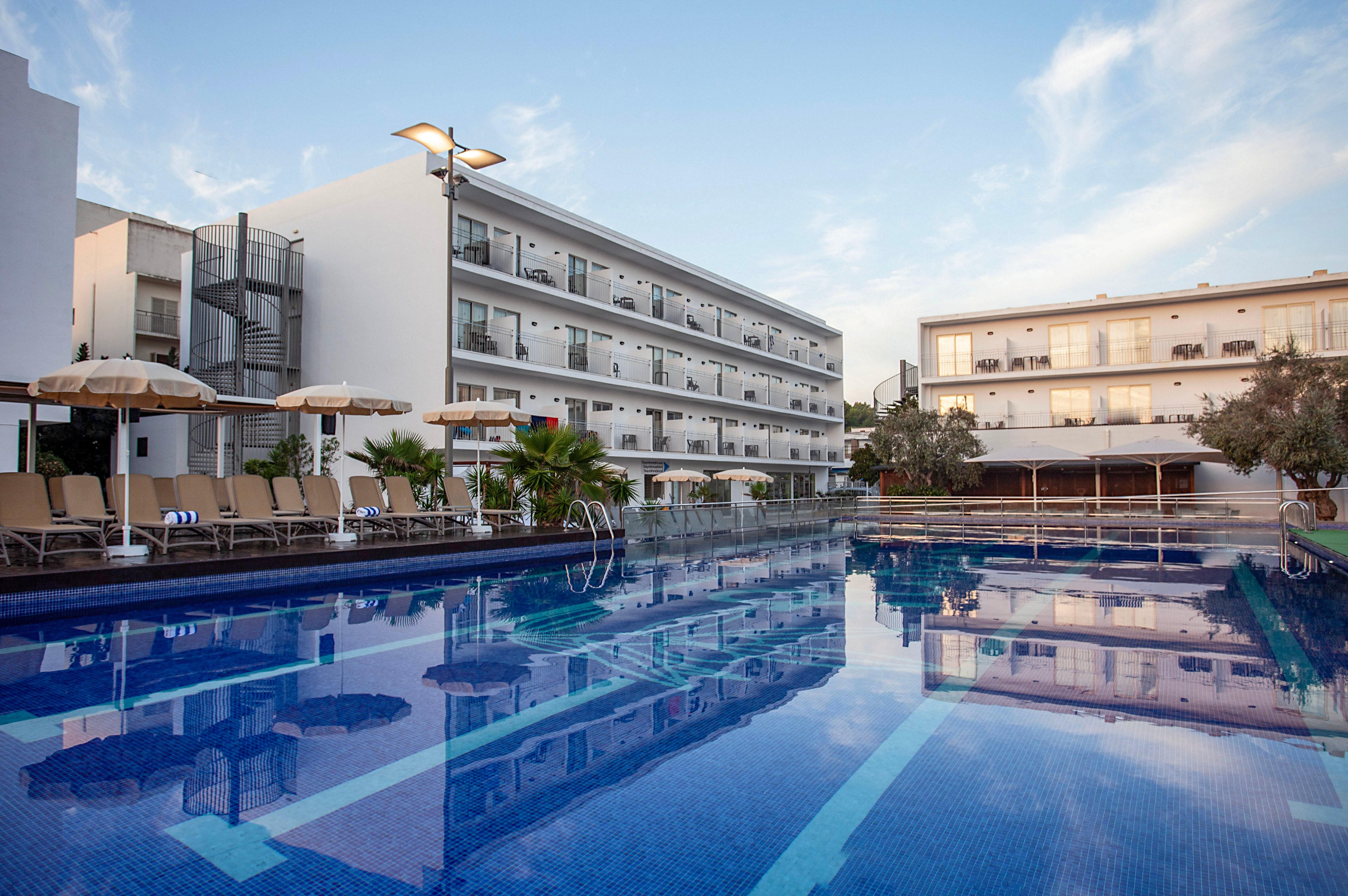A large outdoor swimming pool reflects the modern hotel buildings surrounding it. Sun loungers and beige parasols line the poolside, suggesting a relaxing vacation setting. The multi-story white structures feature balconies and external staircases, with soft evening lighting creating a calm atmosphere. Clear blue water and tiled surfaces emphasize the clean, contemporary resort style.