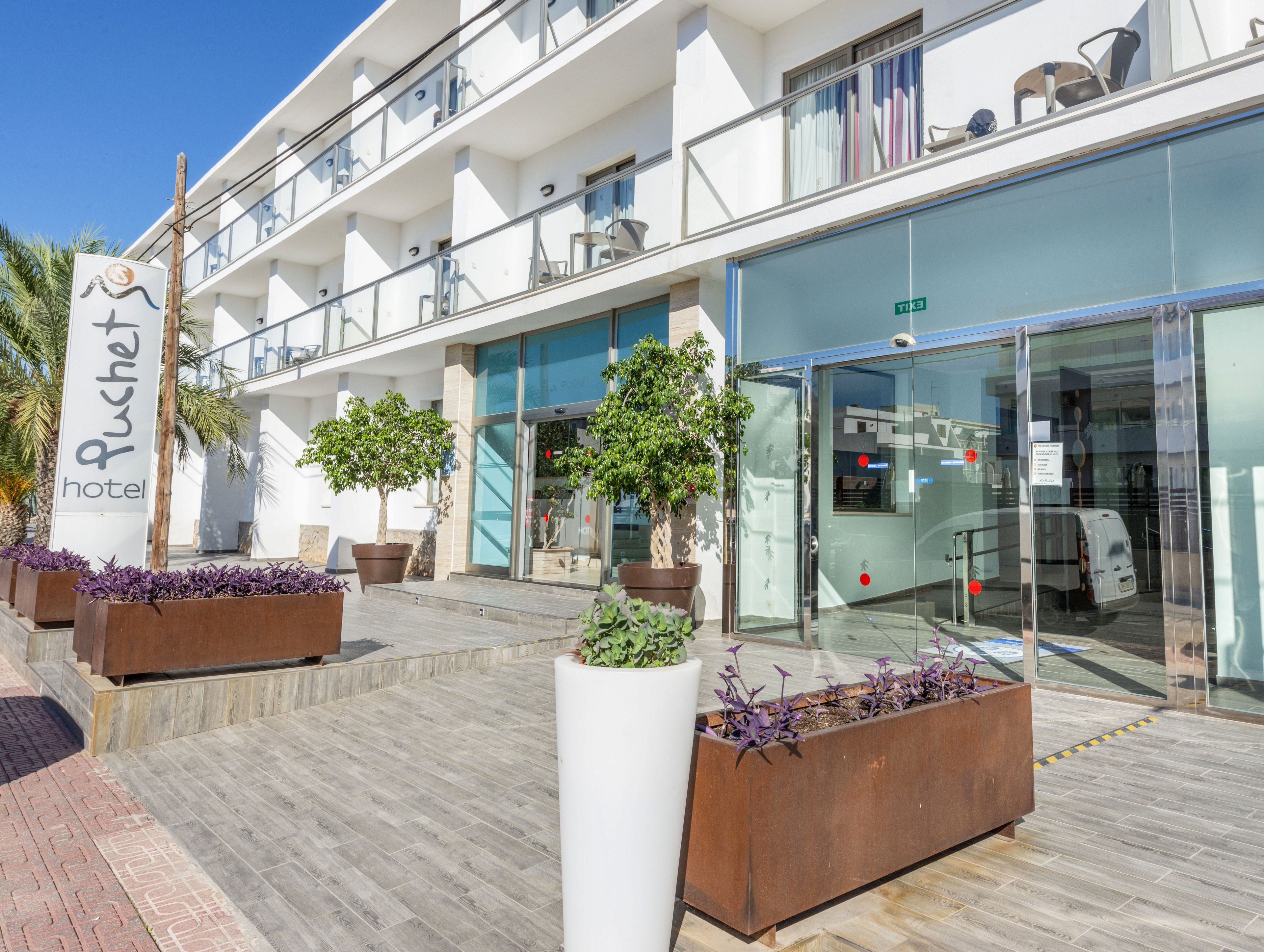 The image shows the exterior entrance of the Puchet hotel on a bright sunny day. A tall sign with the word "Puchet" and "hotel" stands beside a wooden walkway leading to glass doors. Contemporary white architecture with balconies and railings frames the building, while large planters with green trees and purple plants add color. The scene suggests a clean, modern accommodation in a warm, vacation-friendly setting.