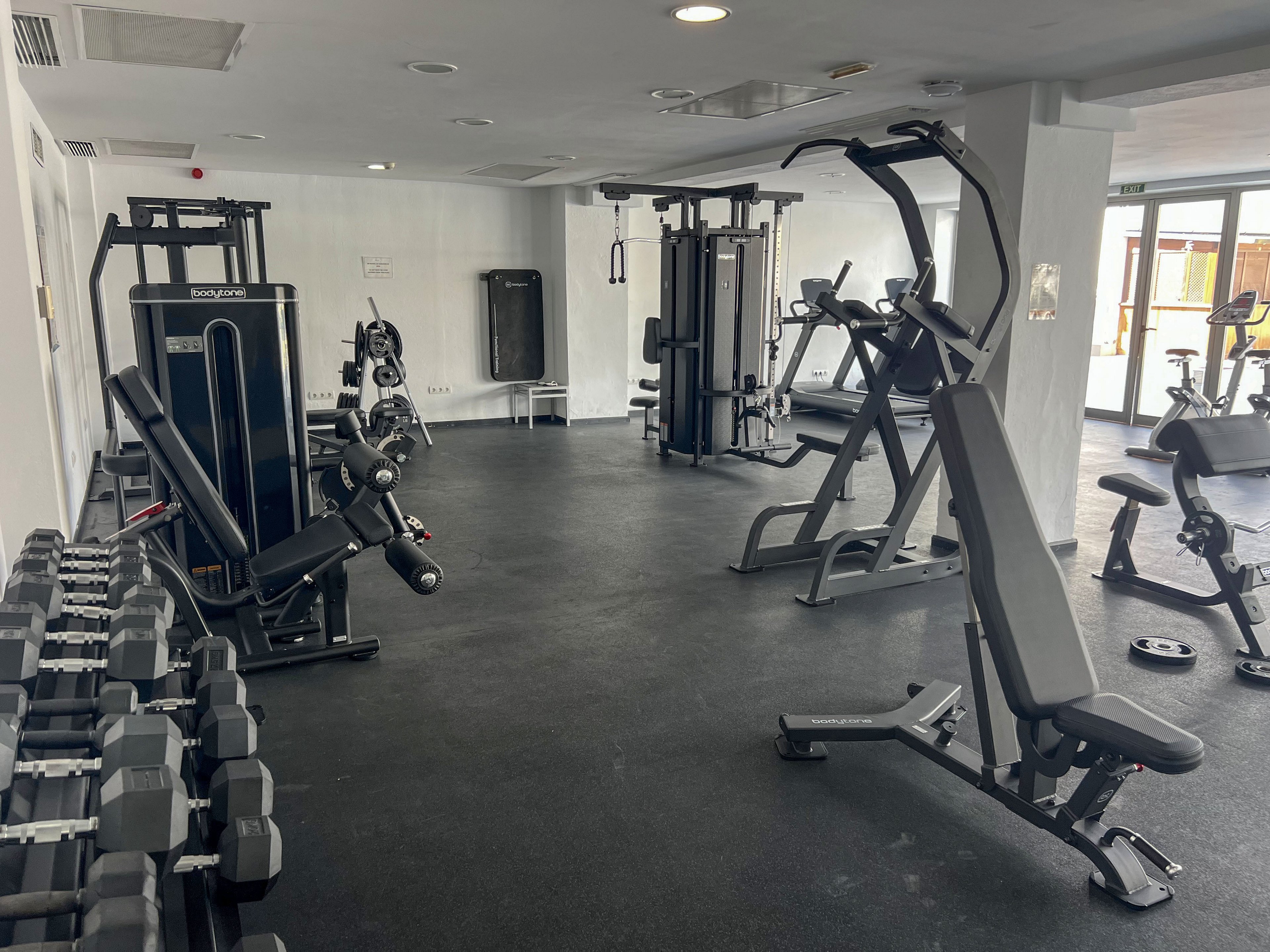 A clean, modern indoor gym features various strength training machines and free weights. Dumbbells are neatly arranged on racks along the left side, while weight machines and a bench press occupy the center. The space has bright white walls, dark flooring, and large windows letting in natural light. No people are present, emphasizing the equipment and open workout area.