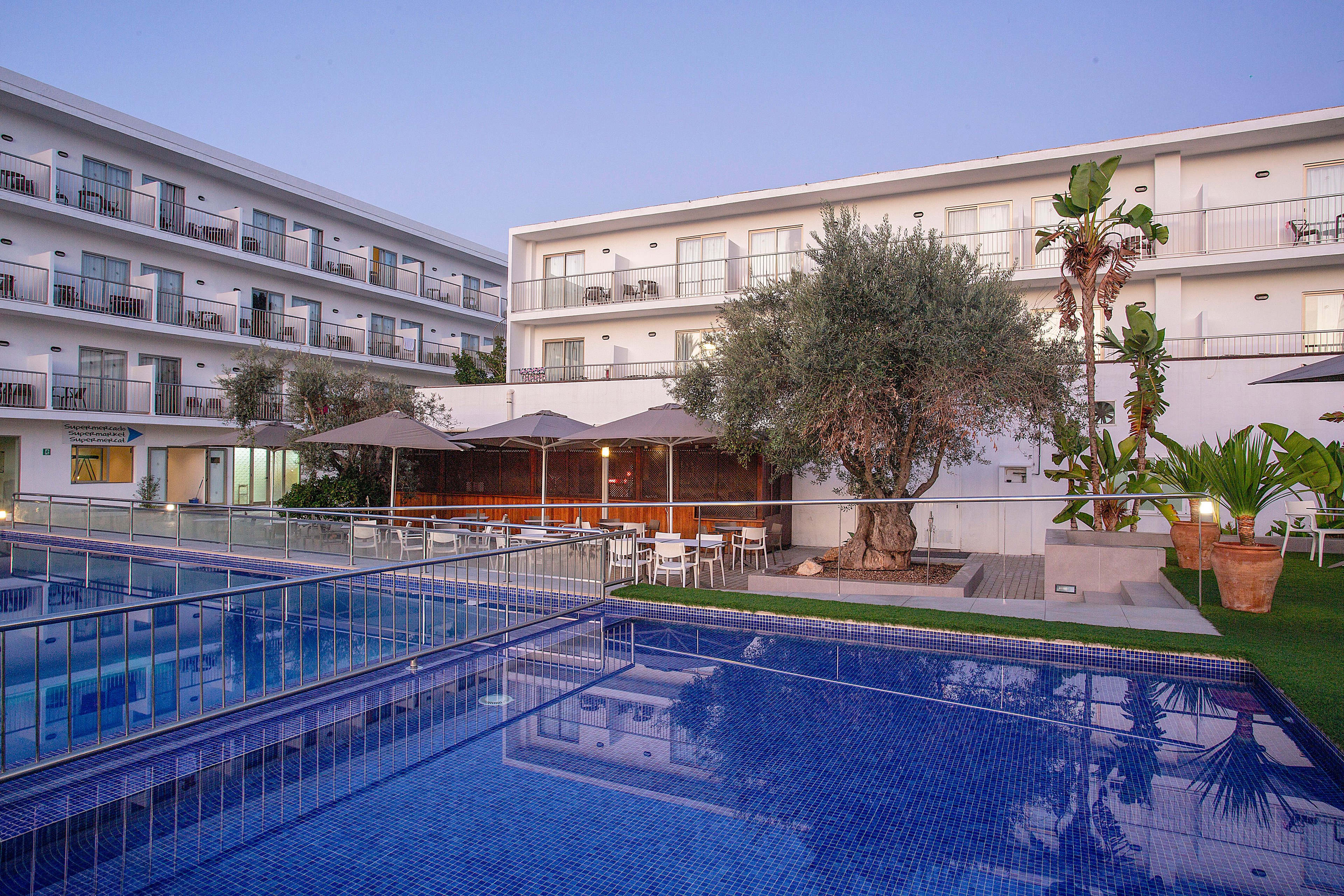 A contemporary hotel complex features multiple stories of rooms with balconies overlooking a large outdoor swimming pool. The scene is captured at dusk, with soft evening light reflecting off the calm blue water. A terrace area with tables, chairs, and umbrellas sits beside a prominent tree, creating a relaxed social space. Palm trees, greenery, and clean architectural lines contribute to a tranquil vacation atmosphere.