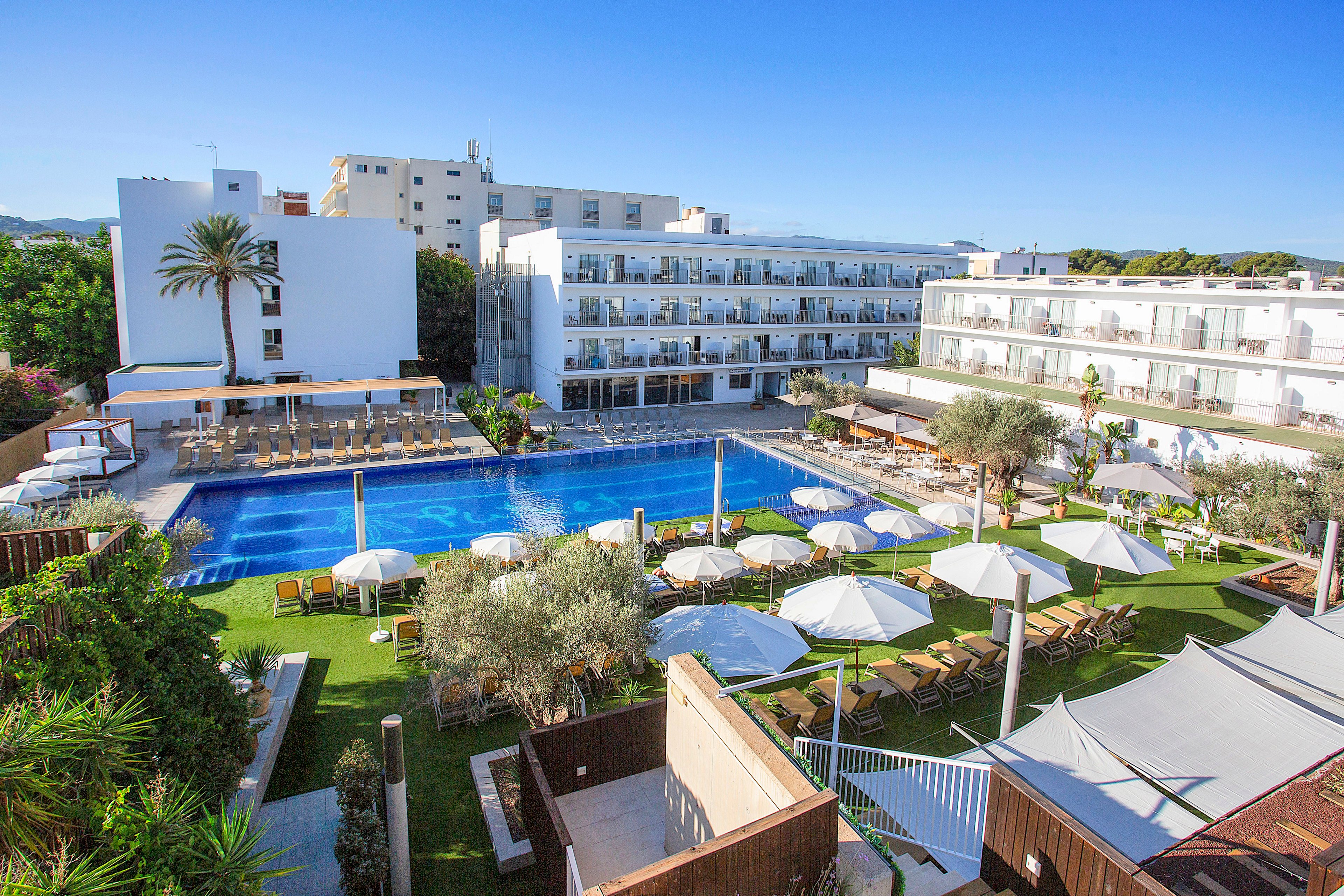 A contemporary resort hotel features a long rectangular outdoor swimming pool surrounded by sun loungers and umbrellas. Multiple white multi-story buildings with balconies overlook the pool area, creating a bright and open courtyard setting. Green lawns, palm trees, and landscaped plants add a tropical feel to the scene. The overall atmosphere is sunny, relaxed, and ideal for vacation leisure.