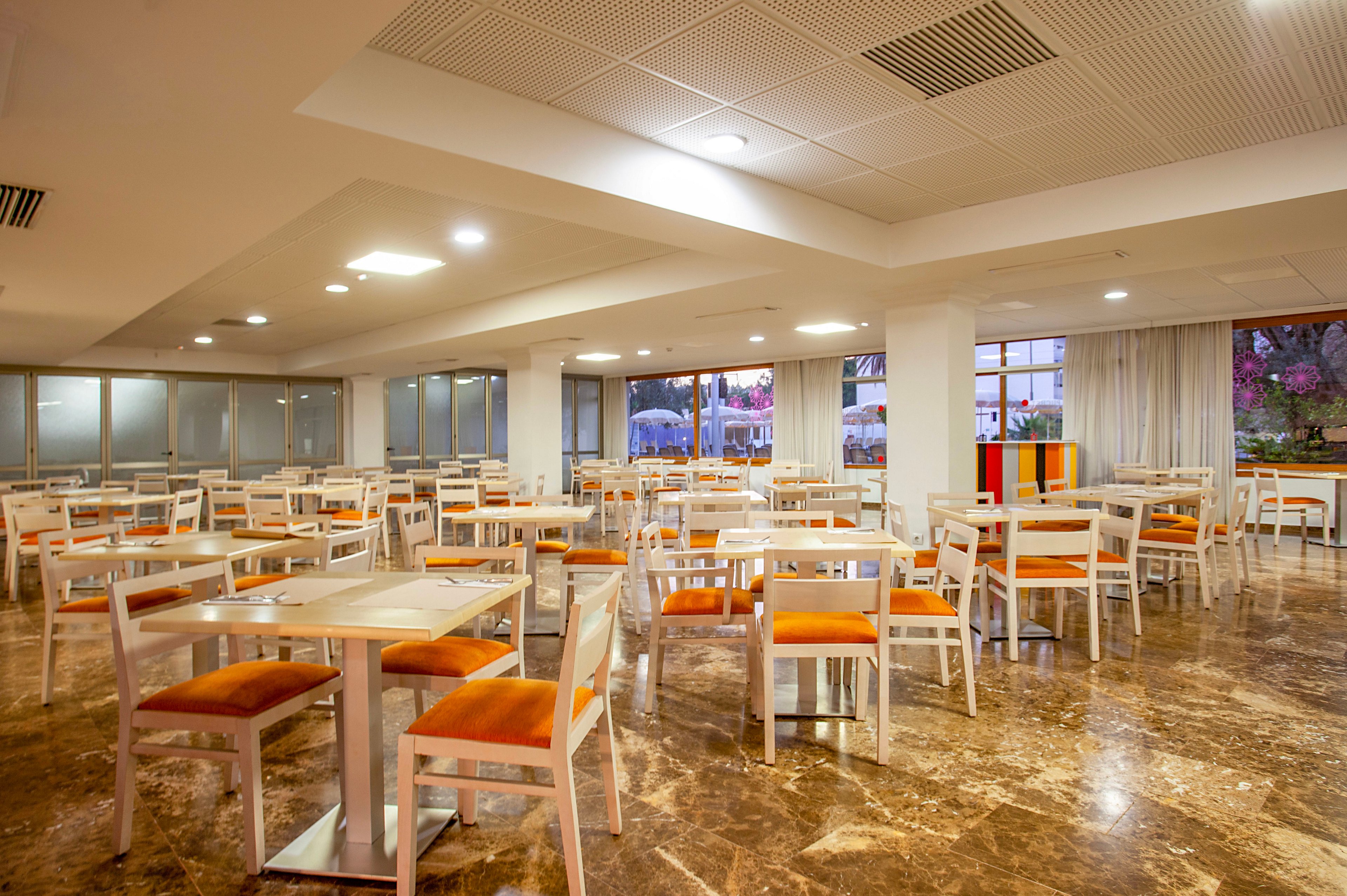 A spacious cafeteria interior features rows of white tables paired with orange cushioned chairs. The room has a polished marble floor and a white ceiling with recessed lighting and ventilation panels. Large windows and glass doors line the walls, revealing greenery and colorful outdoor elements. The overall atmosphere is clean, bright, and ready for guests.