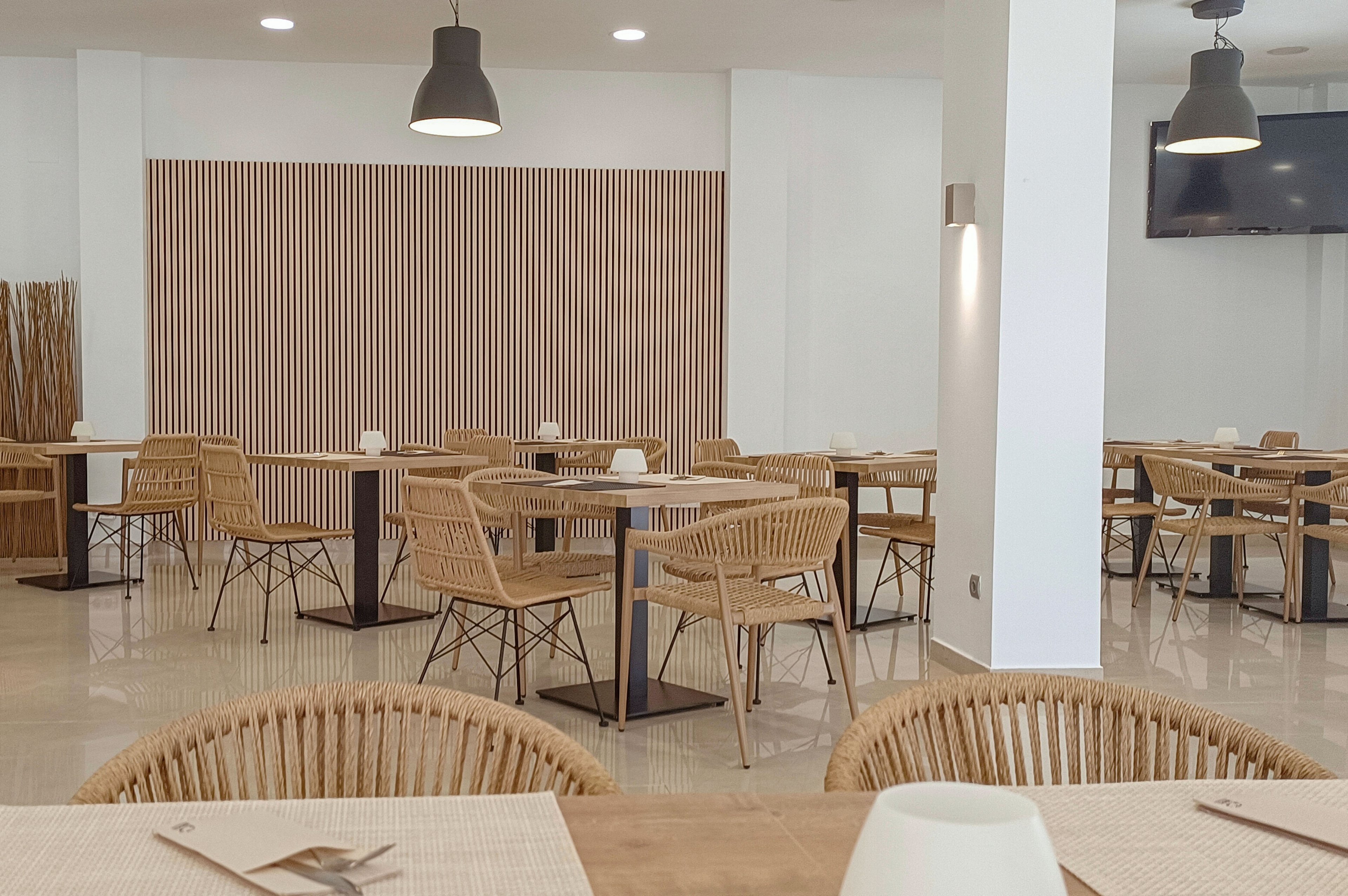 Contemporary dining area featuring light wicker chairs and simple square tables. The setting is clean and minimalist, with neutral tones and a striped wooden wall panel as a focal point. Ceiling spotlights and pendant lamps provide soft, even lighting across the room. A television screen is mounted on the wall, adding a modern touch to the calm restaurant interior.
