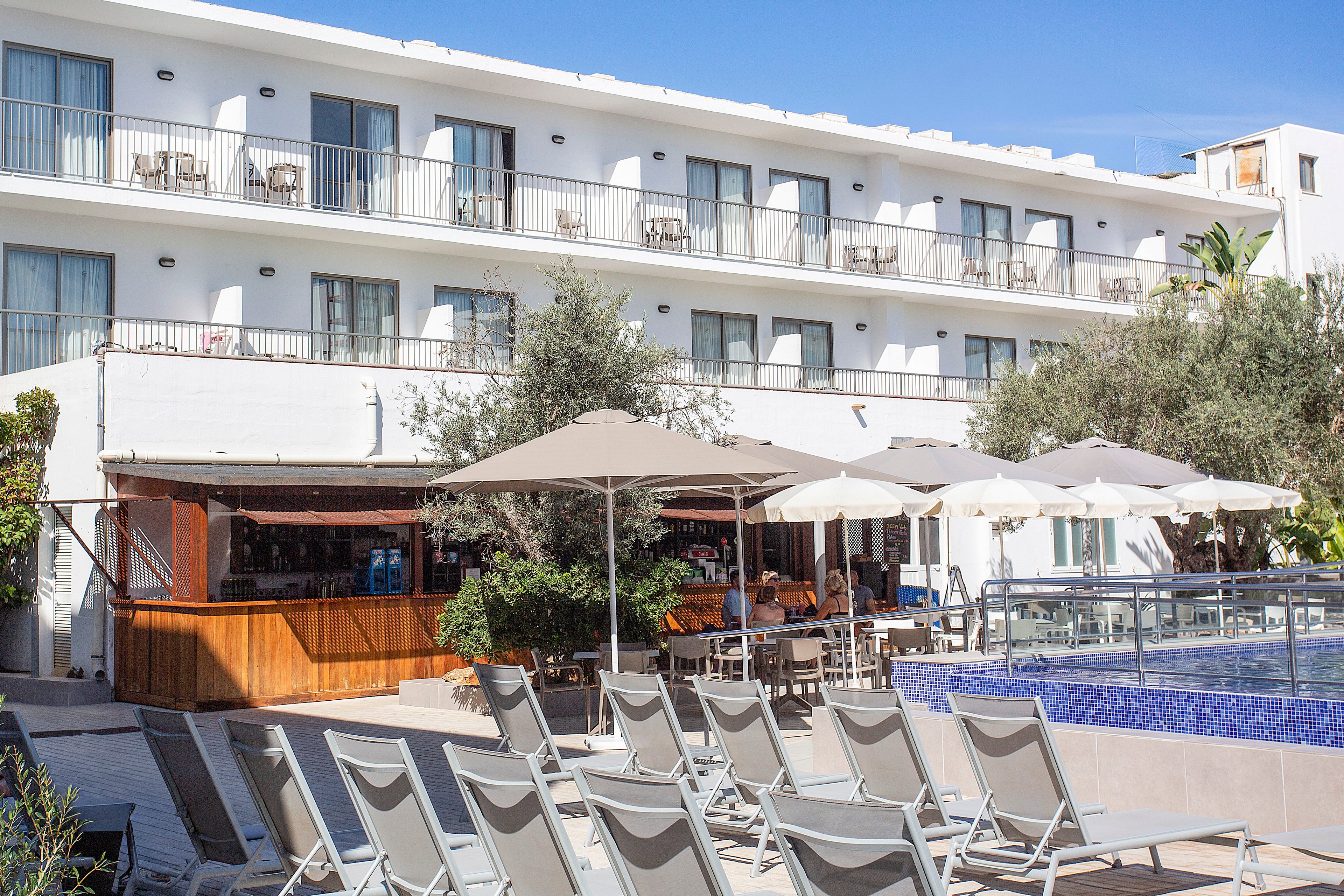 A contemporary white hotel building overlooks a sunny outdoor pool area lined with gray sun loungers. Large umbrellas shade a poolside bar and seating area, creating a relaxed vacation atmosphere. Glass railings border the blue-tiled pool, while balconies with sliding doors run along the upper floors. The scene suggests a warm, resort-style setting ideal for leisure and relaxation.