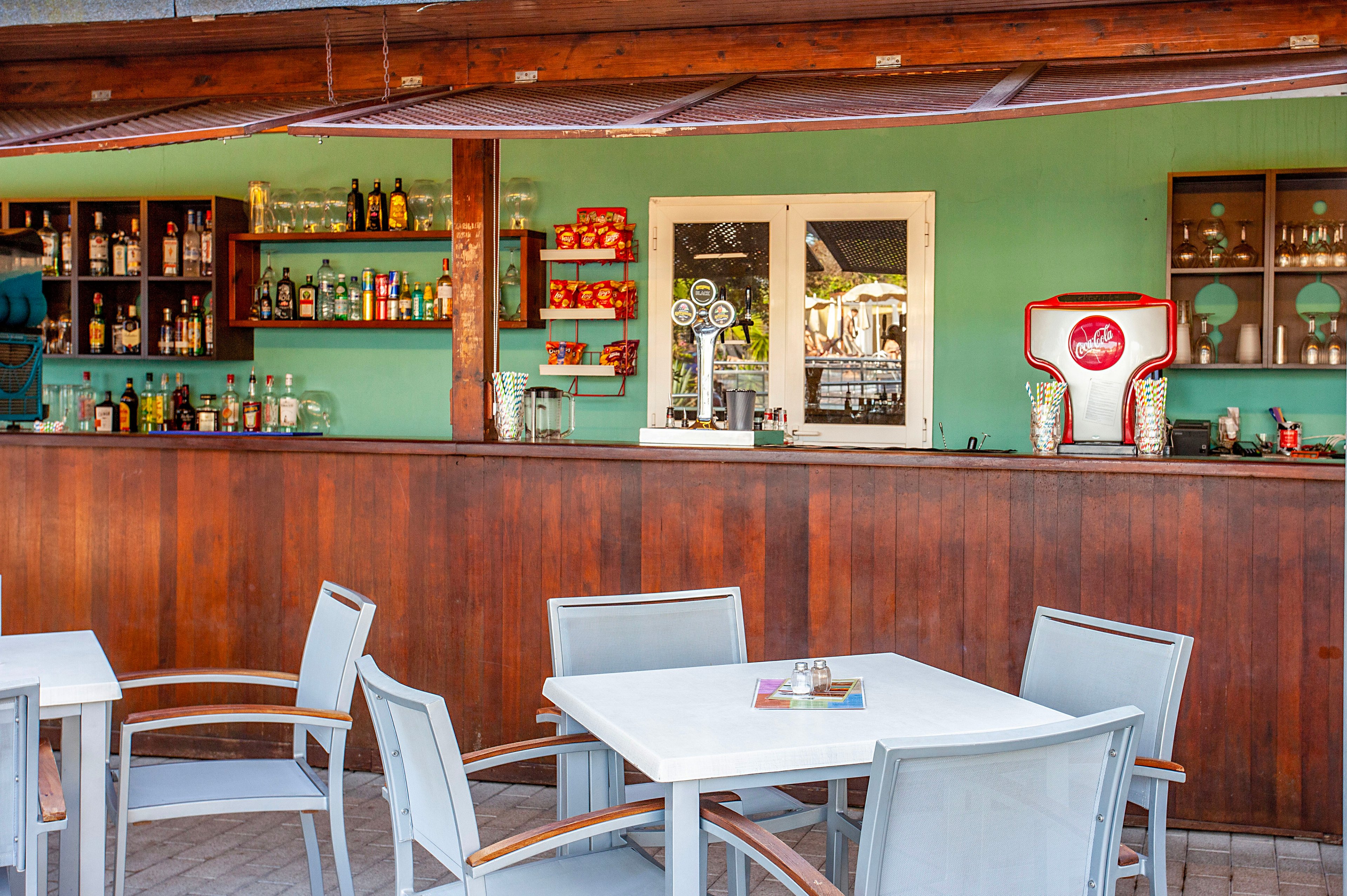An open-air bar features a long wooden counter with bottles of liquor and beverages neatly arranged on wall shelves. Light gray tables and chairs are set in front of the bar, suggesting a casual outdoor seating area. A turquoise wall, glassware, and a red and white drink dispenser add bright accents to the scene. The overall atmosphere feels relaxed and inviting, suitable for vacation or resort leisure.