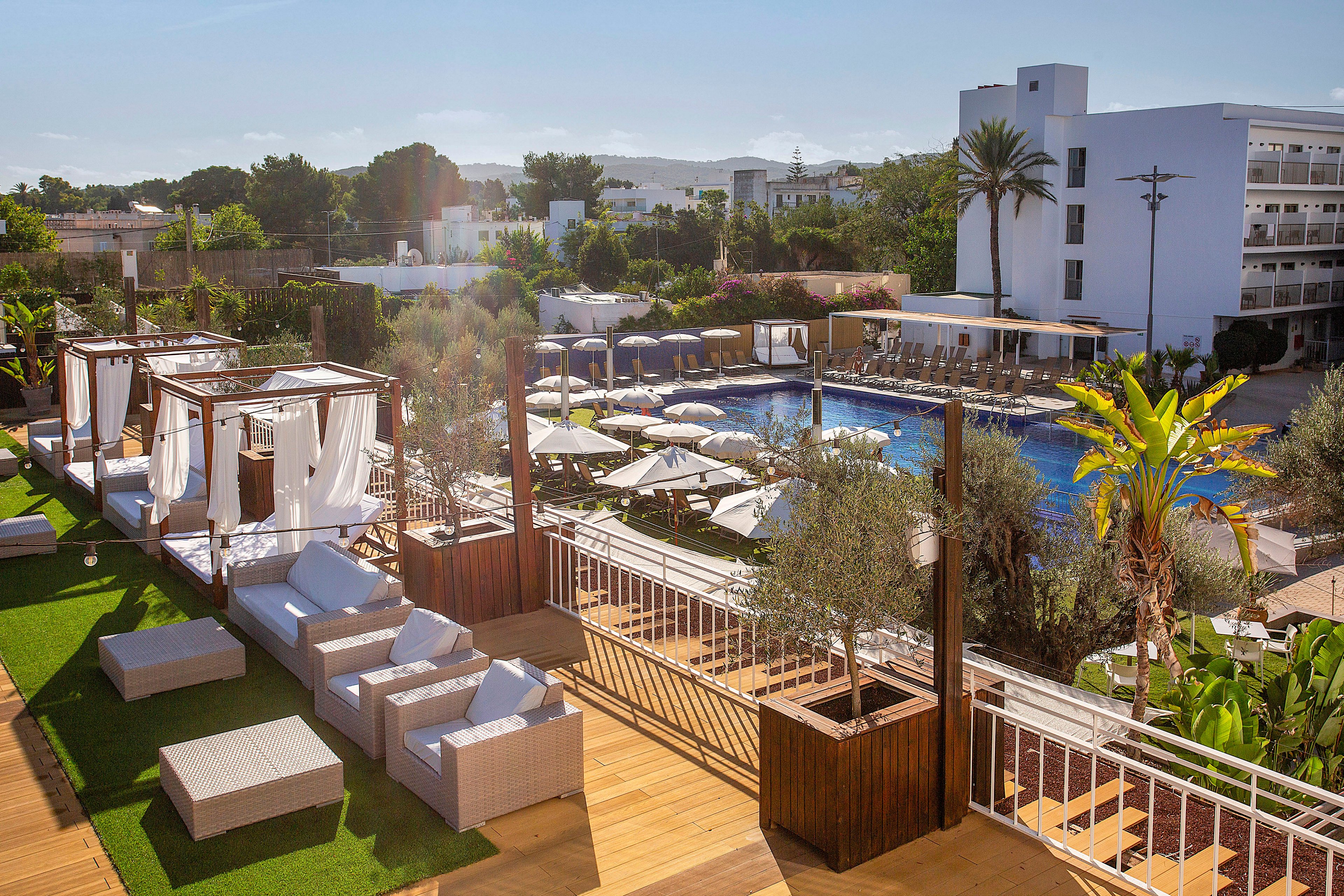 An elevated terrace overlooks a resort swimming pool surrounded by sun loungers and umbrellas. The foreground features modern outdoor seating and canopy cabanas arranged on wooden decking and artificial grass. Palm trees and lush greenery frame the pool area, with white hotel buildings in the background under a clear blue sky. The scene suggests a relaxed vacation atmosphere in a warm, sunny destination.