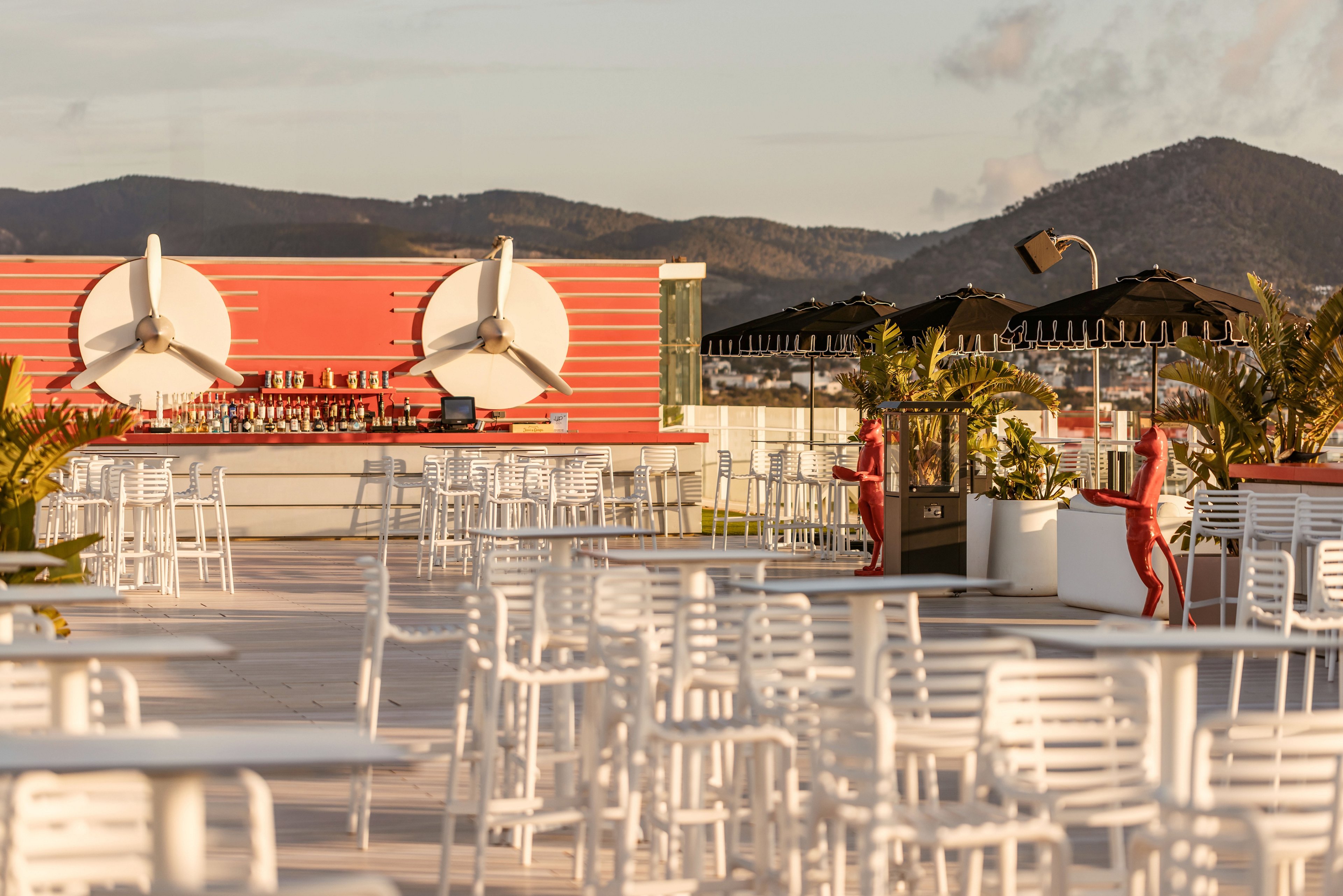 An open-air rooftop bar features rows of white tables and chairs under warm sunlight. A long red bar counter with large circular decor elements stands in the background. Black umbrellas, potted palms, and distant hills create a relaxed, scenic atmosphere. The space appears ready for guests, emphasizing leisure and outdoor dining.