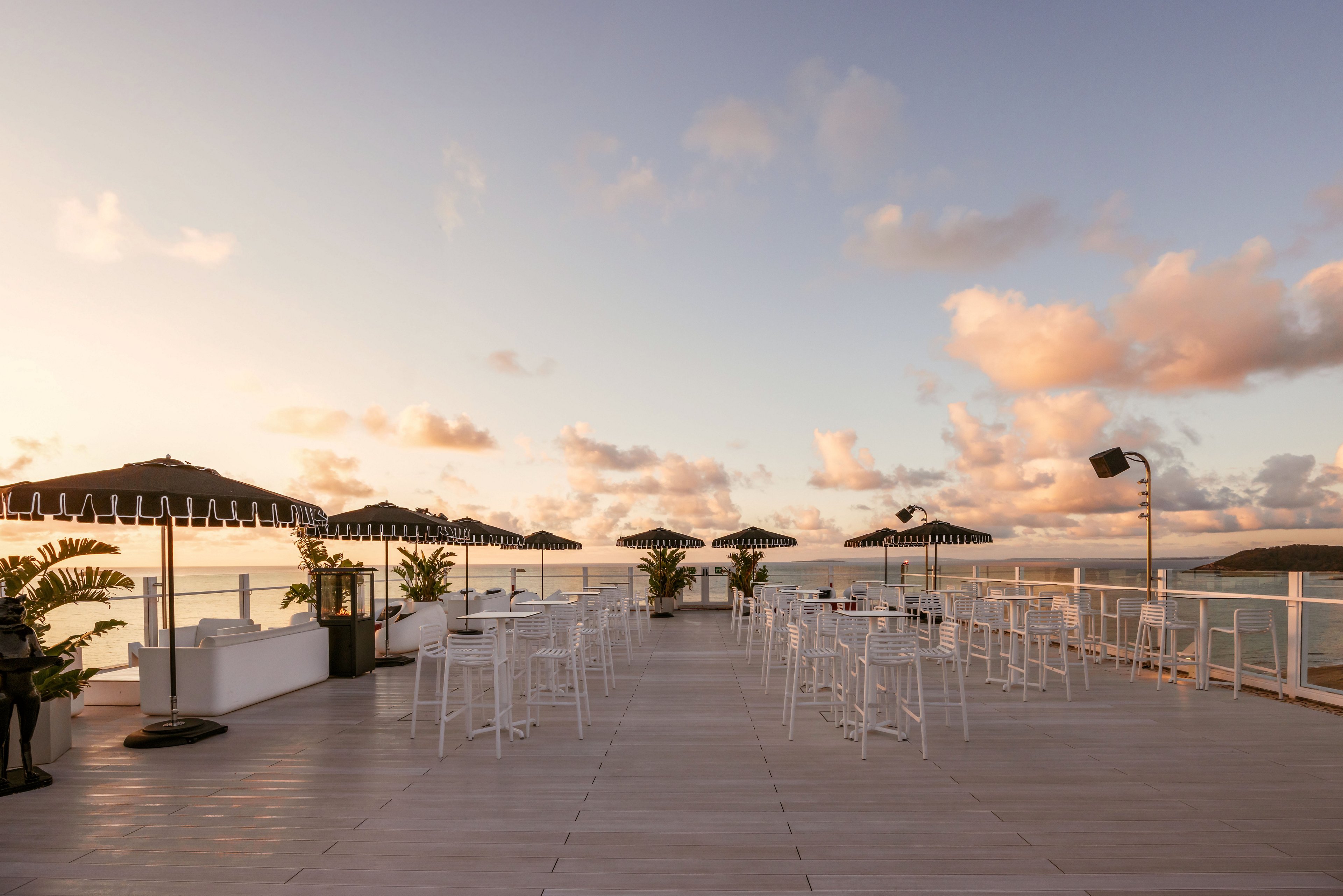 An open-air rooftop terrace is set with white tables, chairs, and dark parasols facing a calm ocean view. The scene is captured at sunset, with warm light reflecting off the water and soft clouds scattered across the sky. Glass railings line the deck, enhancing the expansive coastal panorama. Potted plants add a touch of greenery to the otherwise minimalist, modern setting.