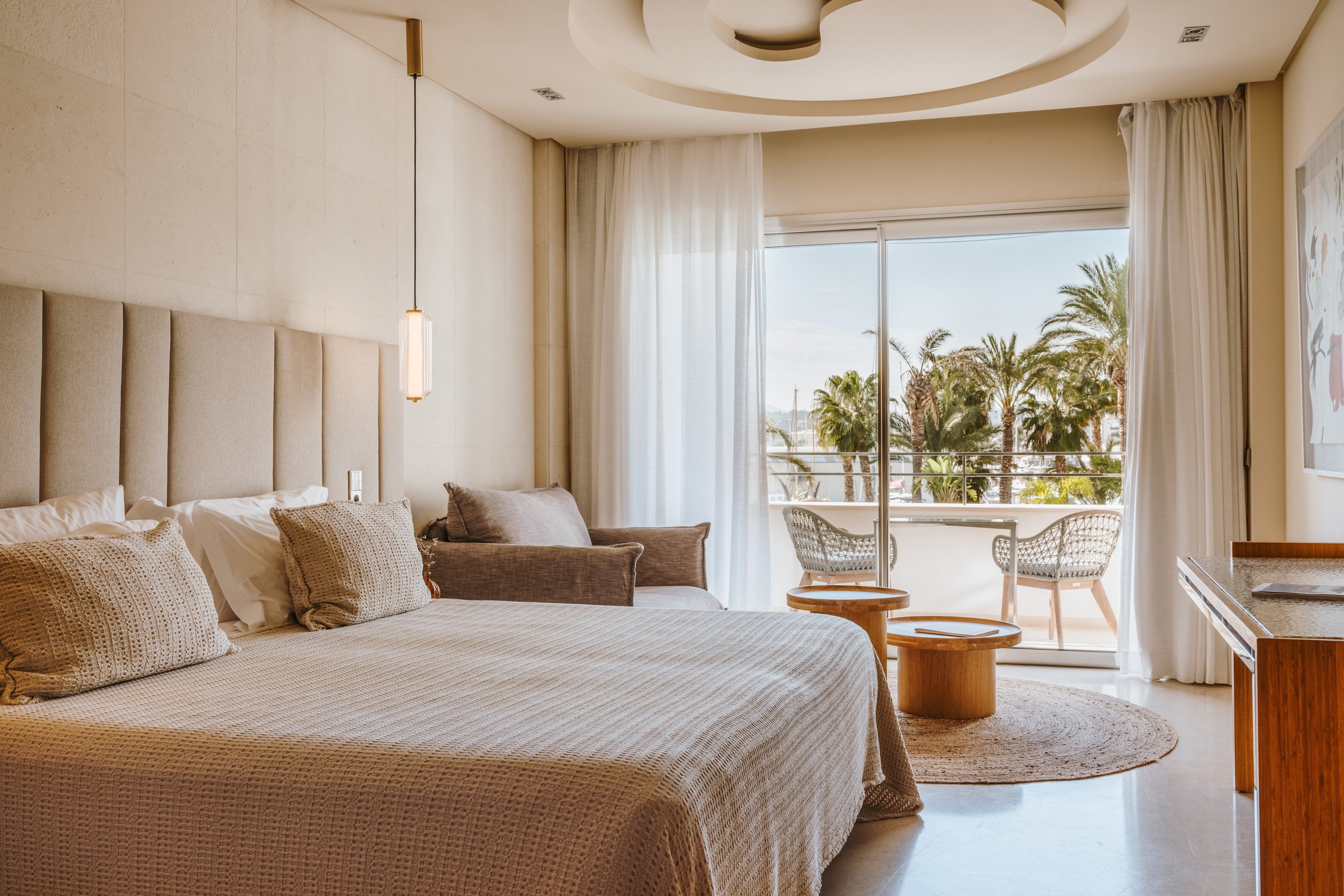 A spacious and elegantly designed hotel room featuring a large bed with neutral-toned bedding. The room includes a seating area and opens to a balcony with outdoor chairs and a table, overlooking palm trees and a sunny landscape. Warm lighting and soft textures create a cozy and inviting atmosphere.