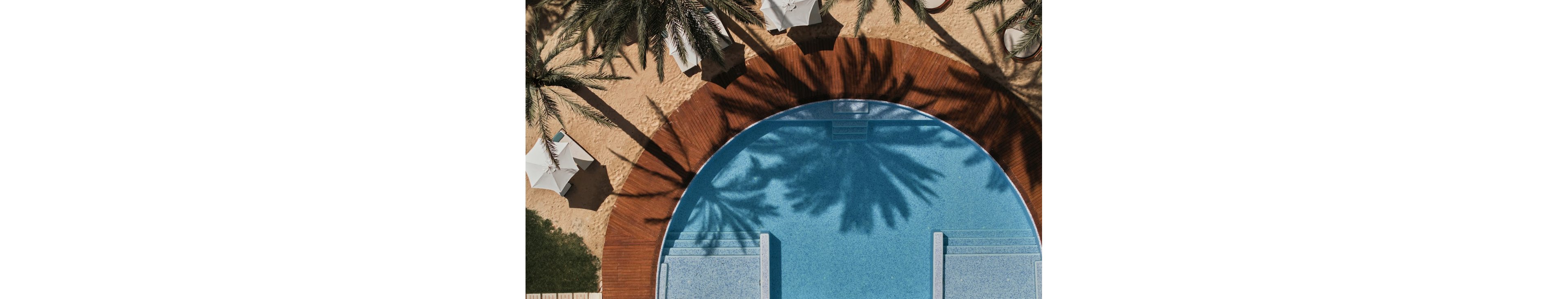 A circular swimming pool is seen from above, bordered by a curved wooden deck and sandy ground. Tall palm trees cast dramatic shadows across the clear blue water and surrounding area. White sun umbrellas and lounge spaces suggest a relaxing resort environment. The composition emphasizes geometric shapes, warm sand tones, and cool aqua hues for a tranquil vacation mood.