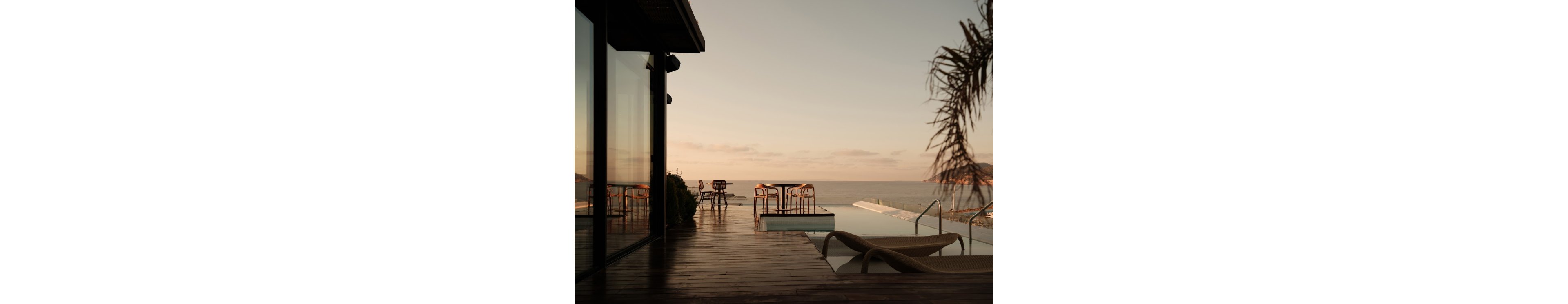A contemporary outdoor terrace overlooks an infinity pool facing the ocean at sunset. Wooden decking leads to lounge chairs and a small seating area with tables and chairs near the water. Soft golden light reflects off the glass windows of the building and the calm sea in the background. The tranquil scene suggests a luxurious coastal retreat with minimal, modern design.