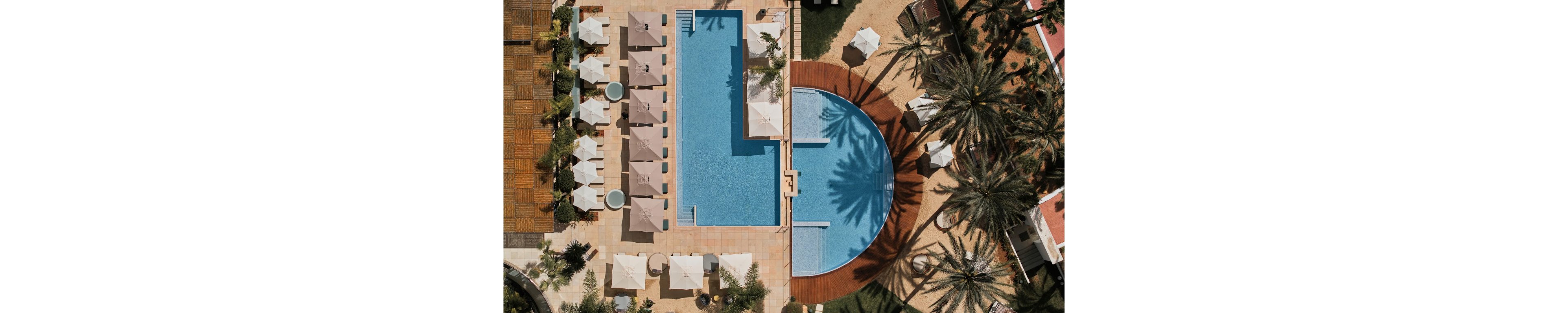 A geometric pair of swimming pools is seen from above in a tropical resort setting. White sun umbrellas and lounge chairs line the tiled pool deck, creating orderly rows around the water. Palm trees cast long shadows across the pools and surrounding walkways, adding texture and contrast. The scene suggests a sunny, relaxing vacation atmosphere with clean, modern design elements.