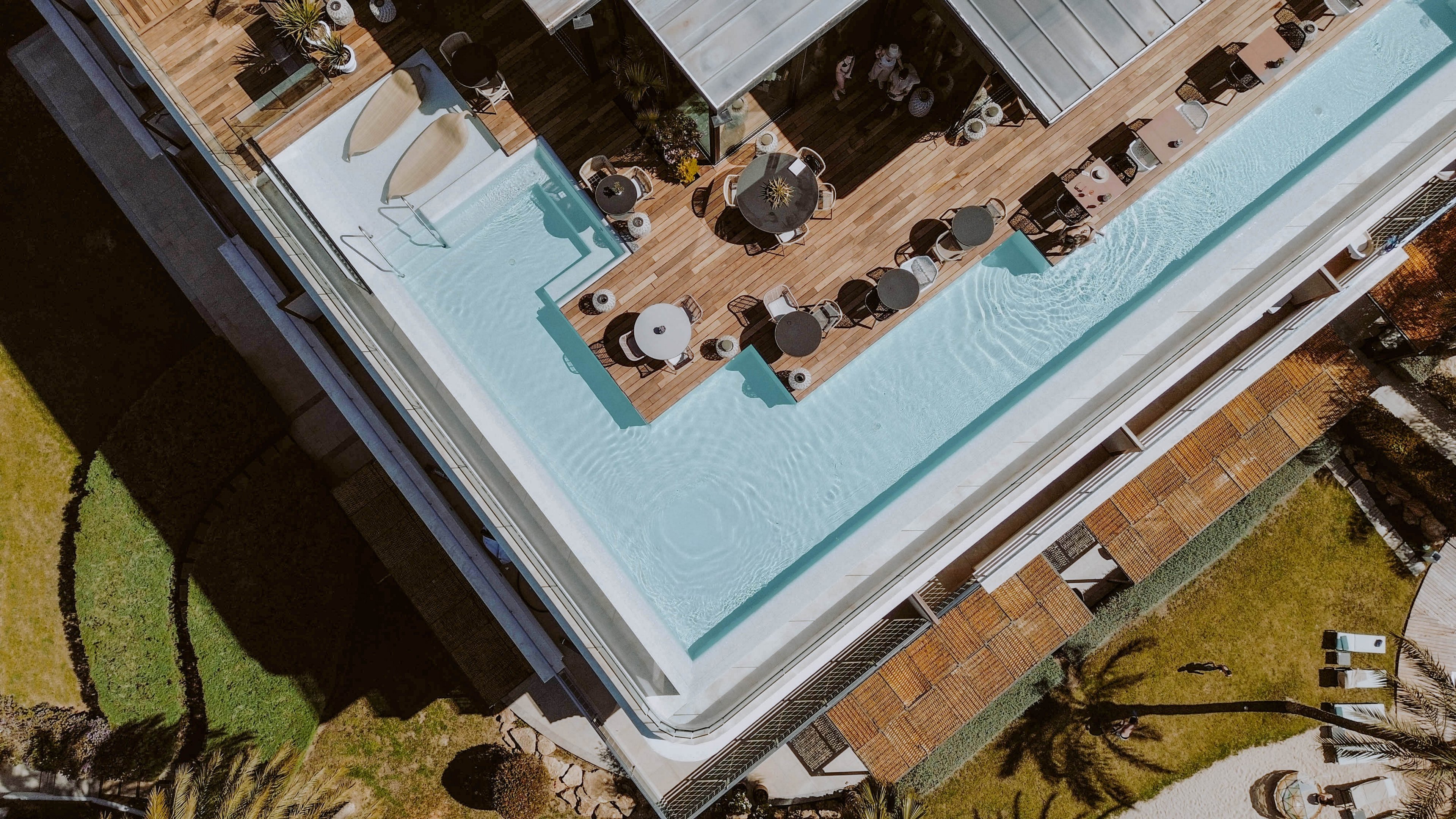This image shows a modern rooftop pool surrounded by a wooden deck with tables, chairs, and umbrellas. A single person is floating in the pool, while others are seated or walking around the lounge area. The setting is bright and sunny, with a relaxed and upscale atmosphere. The pool is rectangular and the surrounding area features landscaped greenery and palm trees.