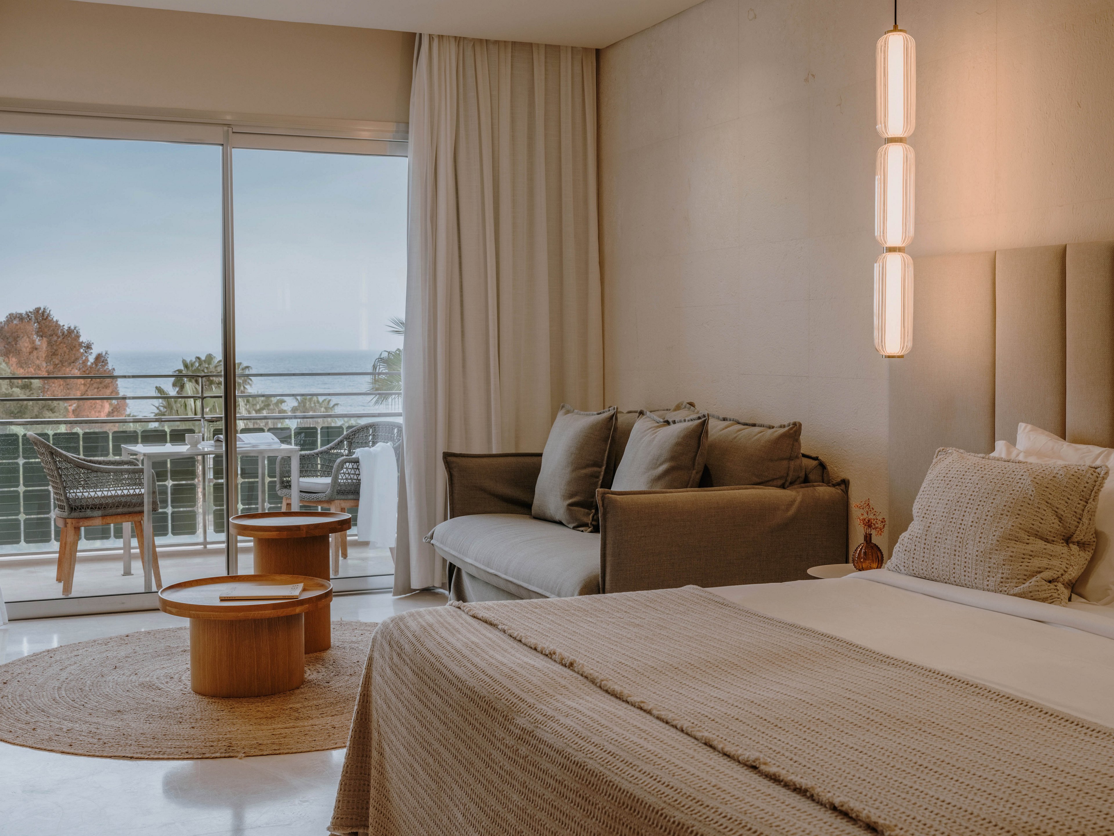 A contemporary hotel room features a large bed with neutral bedding, a gray sofa, and a set of round wooden tables. The space is illuminated by pendant lights and natural light from a sliding glass door leading to a balcony with a scenic ocean view. The room's palette is soft and earthy, creating a calm and inviting atmosphere.