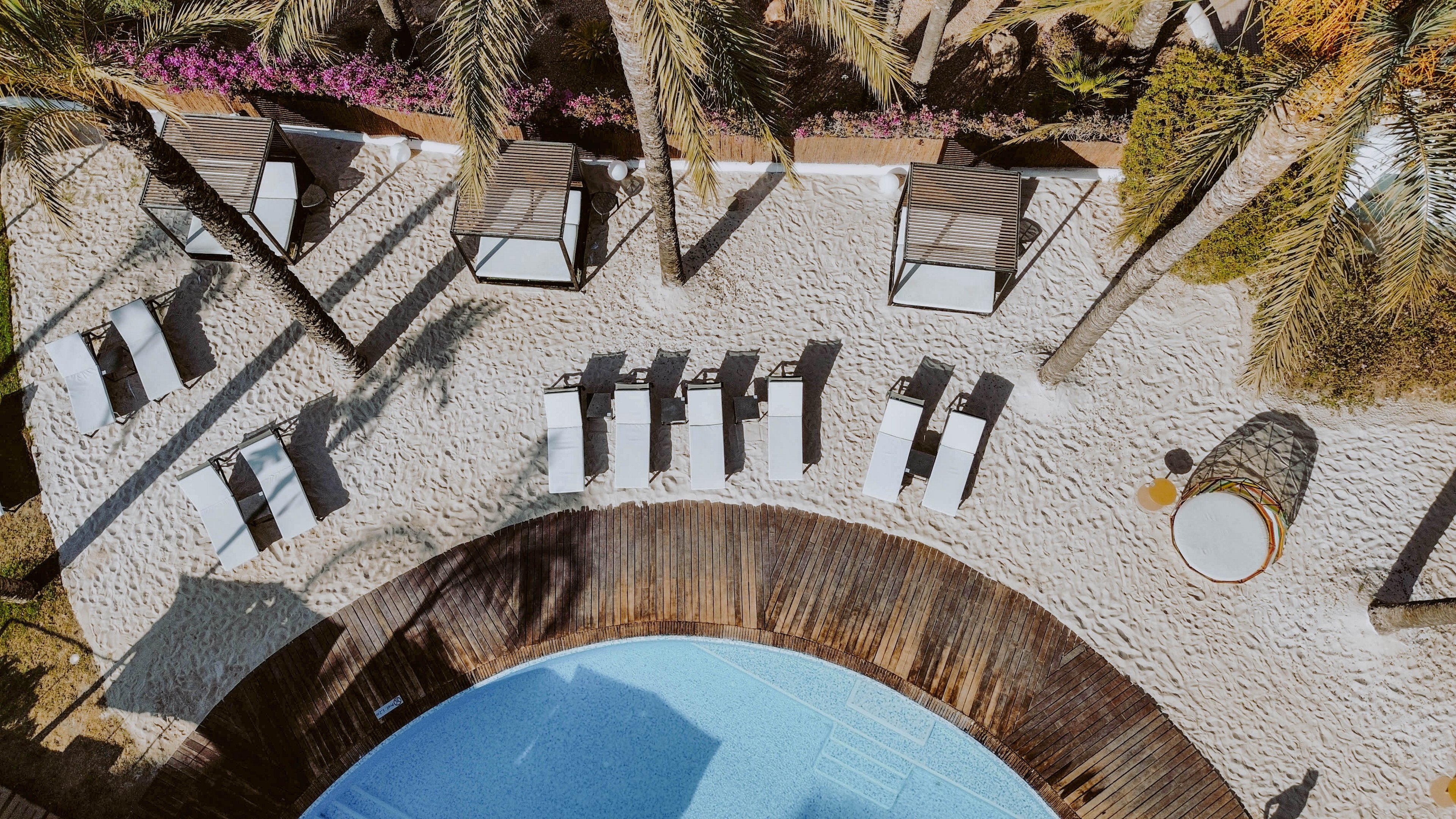 This image shows an overhead perspective of a resort pool area with several white lounge chairs and shaded cabanas arranged on a sandy surface. Palm trees cast shadows across the scene, and a curved wooden deck borders the blue swimming pool. The setting is tranquil and sunlit, evoking a relaxed, tropical vacation mood.