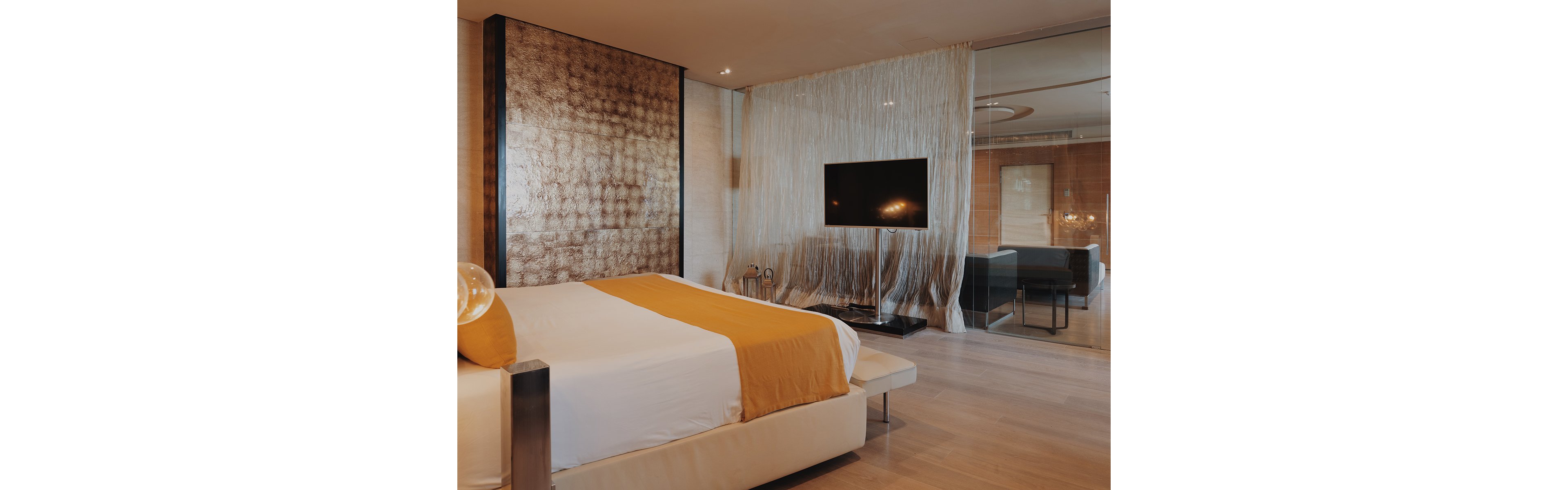 A contemporary hotel bedroom features a large bed with white linens and a mustard yellow throw. The room includes a flat-screen television on a sleek stand positioned in front of a sheer curtain divider. Warm wood flooring and textured metallic wall panels add a sophisticated touch. In the background, a glass partition reveals a separate sitting area with modern furniture and soft ambient lighting.