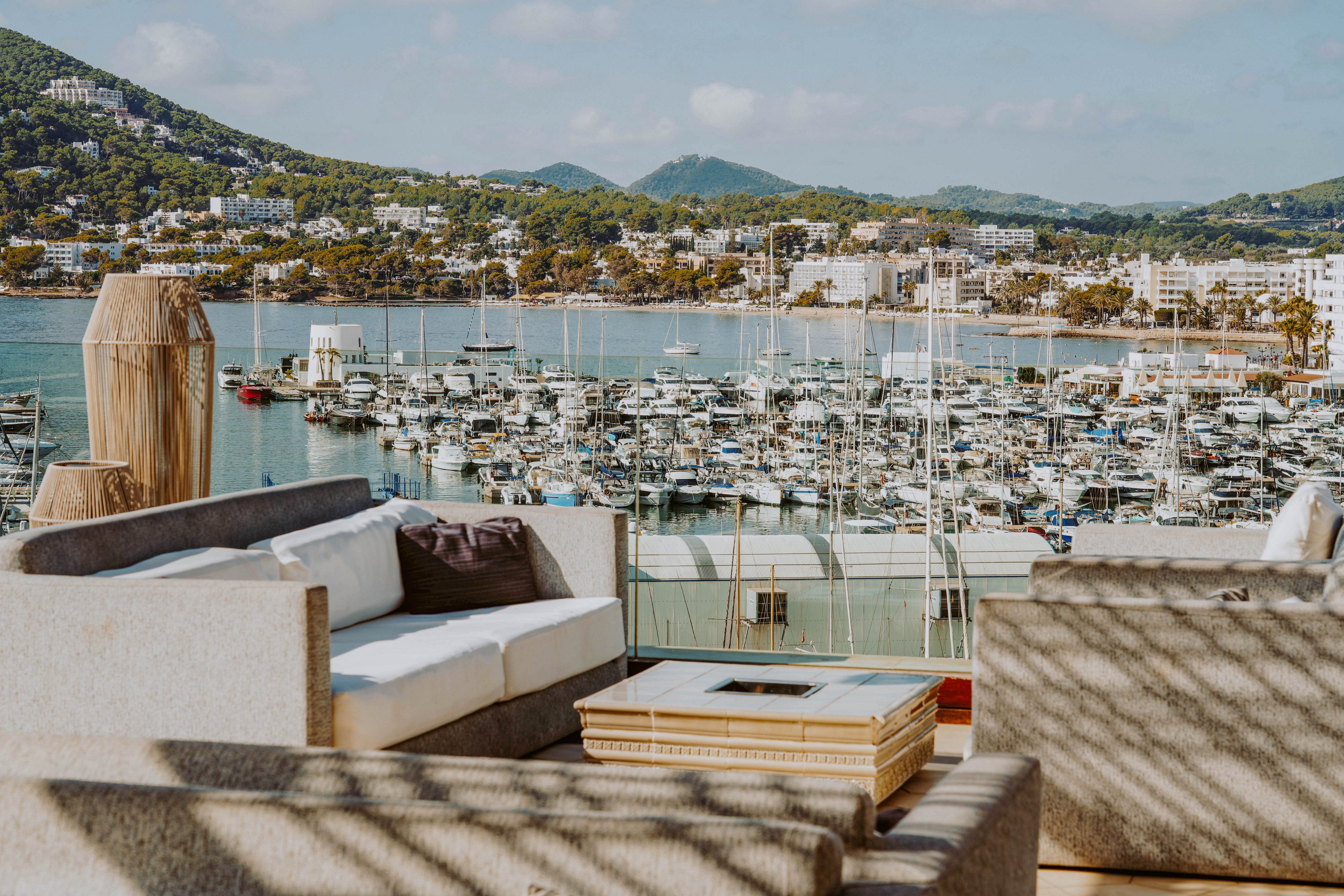 A modern rooftop lounge with cushioned sofas and a low table overlooks a busy marina filled with docked boats. Glass railings frame the view of the harbor and the coastal town beyond. Green hills rise in the background under a bright blue sky with scattered clouds. The scene feels relaxed and inviting, ideal for leisure and waterfront views.