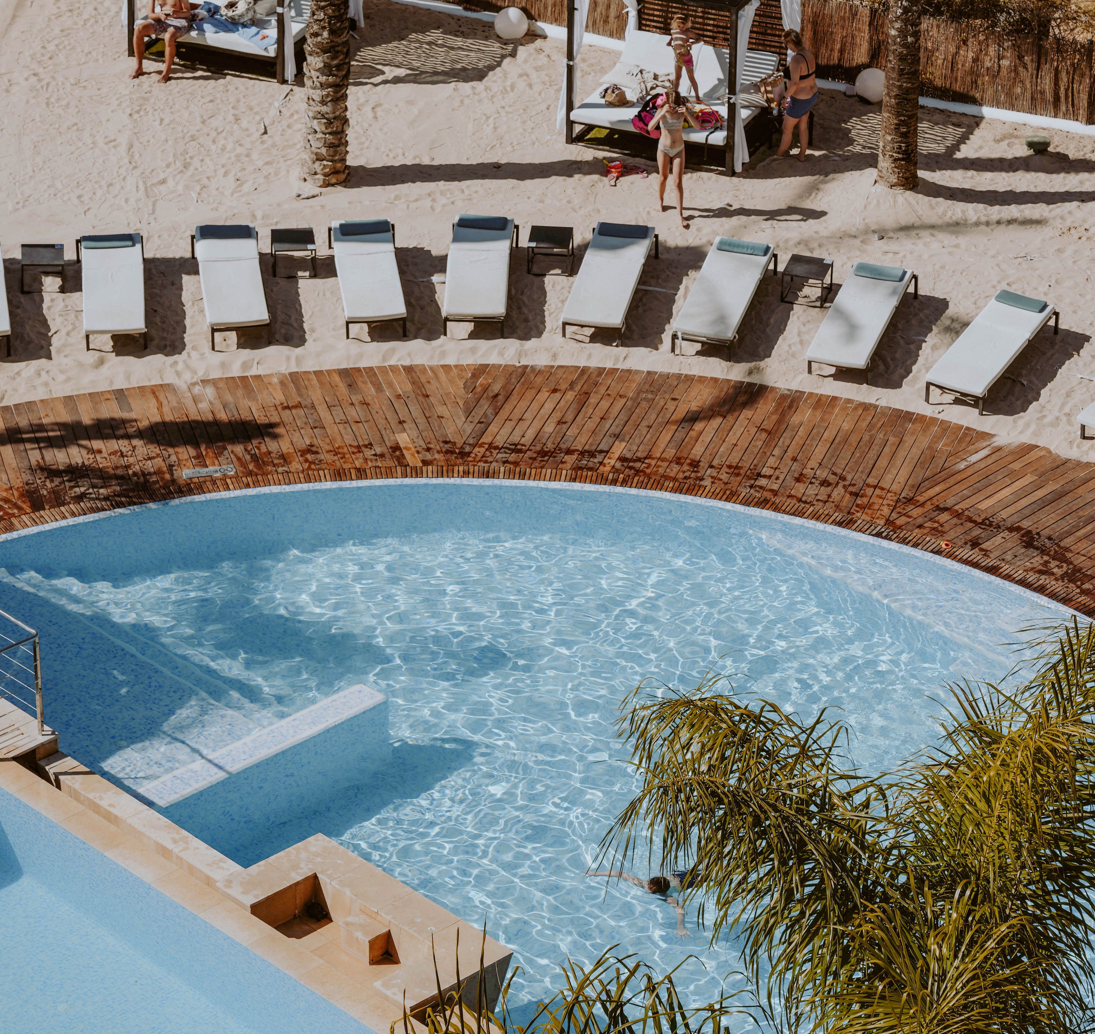 A bright resort swimming pool curves alongside a sandy beach lined with white sun loungers. A wooden deck separates the clear blue water from the beach area, adding warmth and texture to the scene. Palm trees frame the pool, enhancing the tropical vacation atmosphere. A few people relax near cabanas in the background, suggesting a leisurely holiday setting.