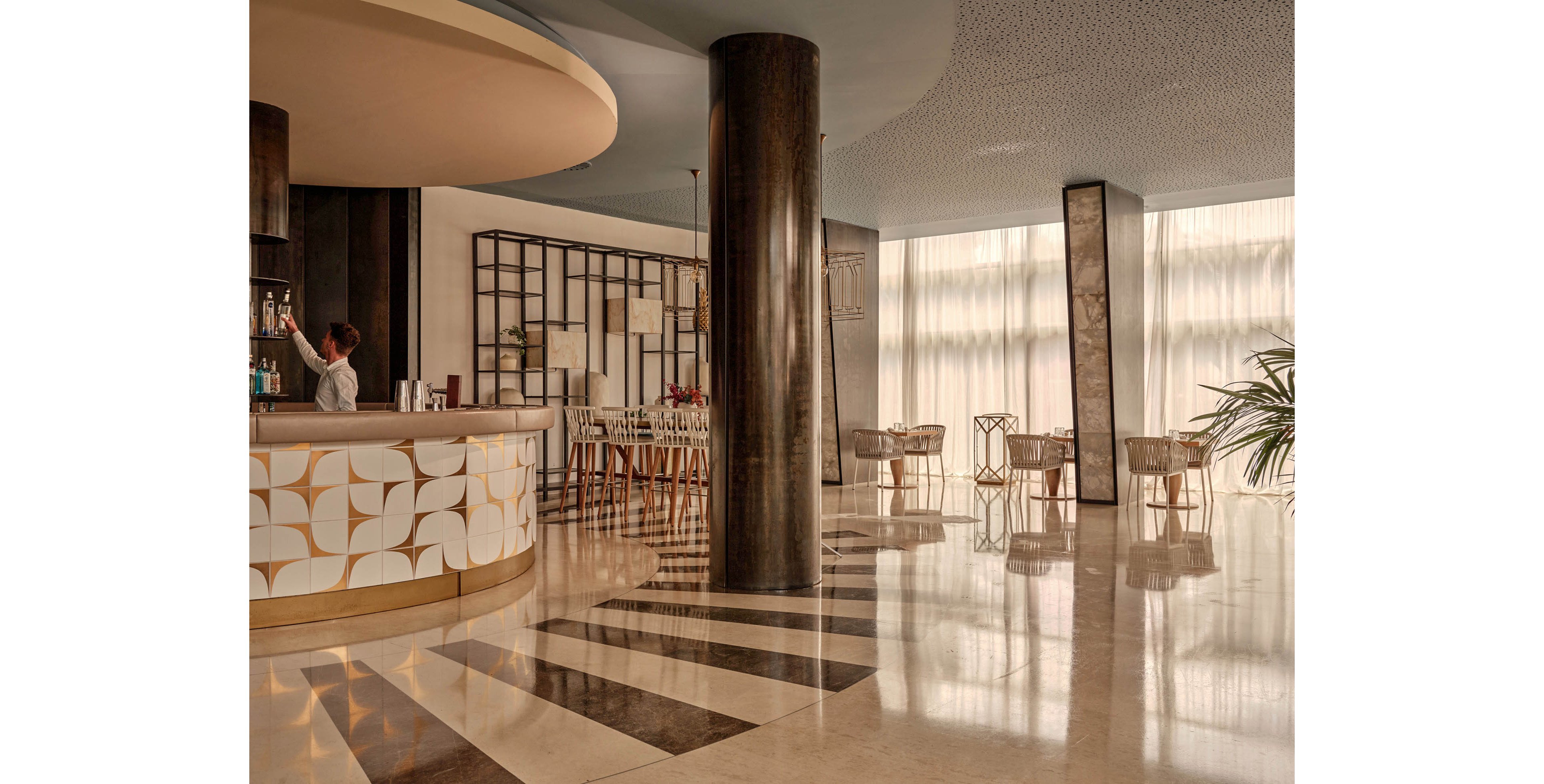 A contemporary hotel lobby bar features a curved counter with geometric patterns and a bartender standing behind it. Tall dark columns and large floor-to-ceiling windows fill the space with soft natural light. Sleek chairs and tables are arranged in the background, creating an inviting seating area. The polished floor reflects the warm tones and clean lines of the modern interior design.