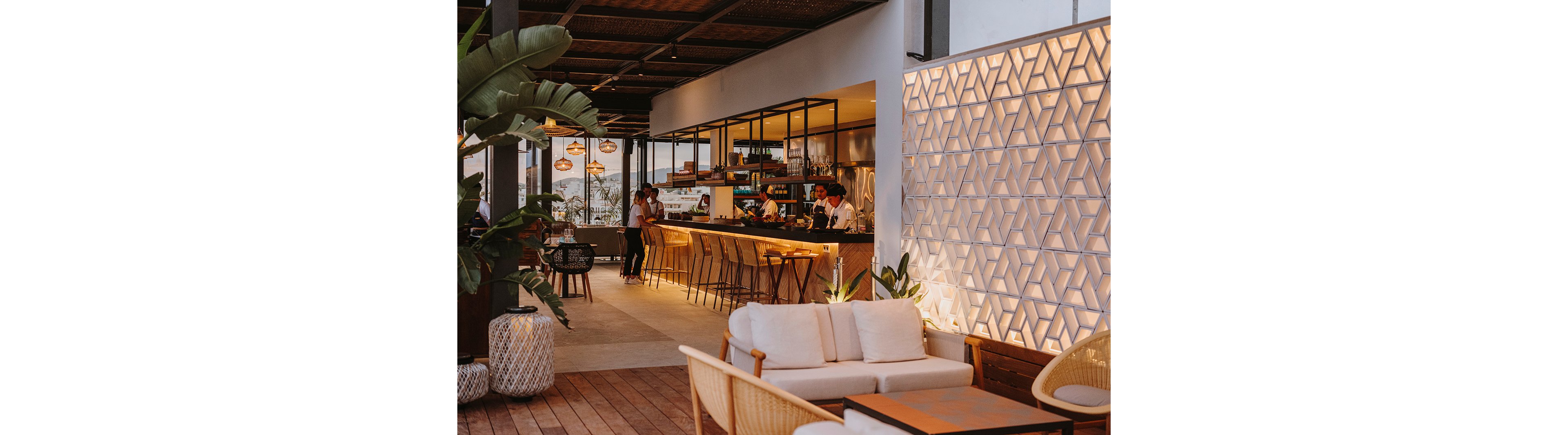 A stylish open-air rooftop bar features a long counter with high stools and warm ambient lighting. Bartenders work behind the bar while guests sit and socialize in the background. Contemporary lounge seating with cushions occupies the foreground, complemented by wooden flooring and decorative plants. A geometric illuminated wall adds texture and a cozy atmosphere to the evening setting.