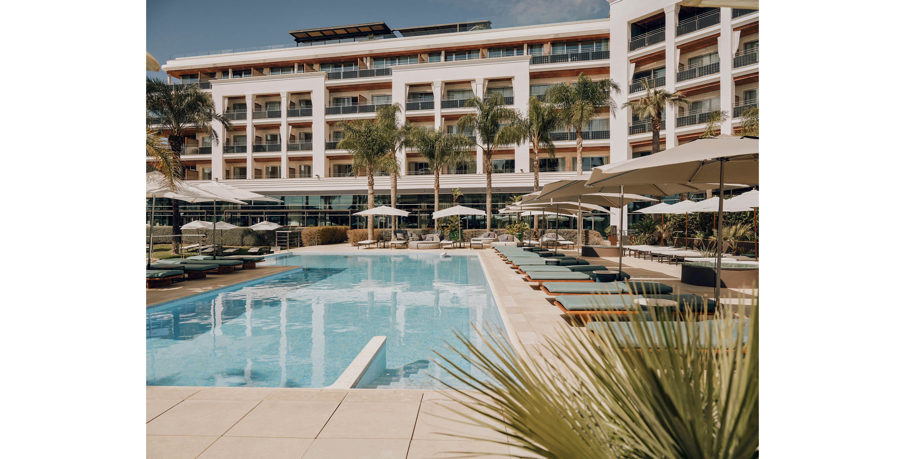 A contemporary multi-story resort hotel features a large outdoor swimming pool in the foreground. Sun loungers with cushions and umbrellas line both sides of the pool, suggesting a relaxing vacation setting. Palm trees and landscaped greenery add a tropical atmosphere around the deck area. The building’s balconies and glass facades reflect the bright daylight, emphasizing a warm, sunny climate.