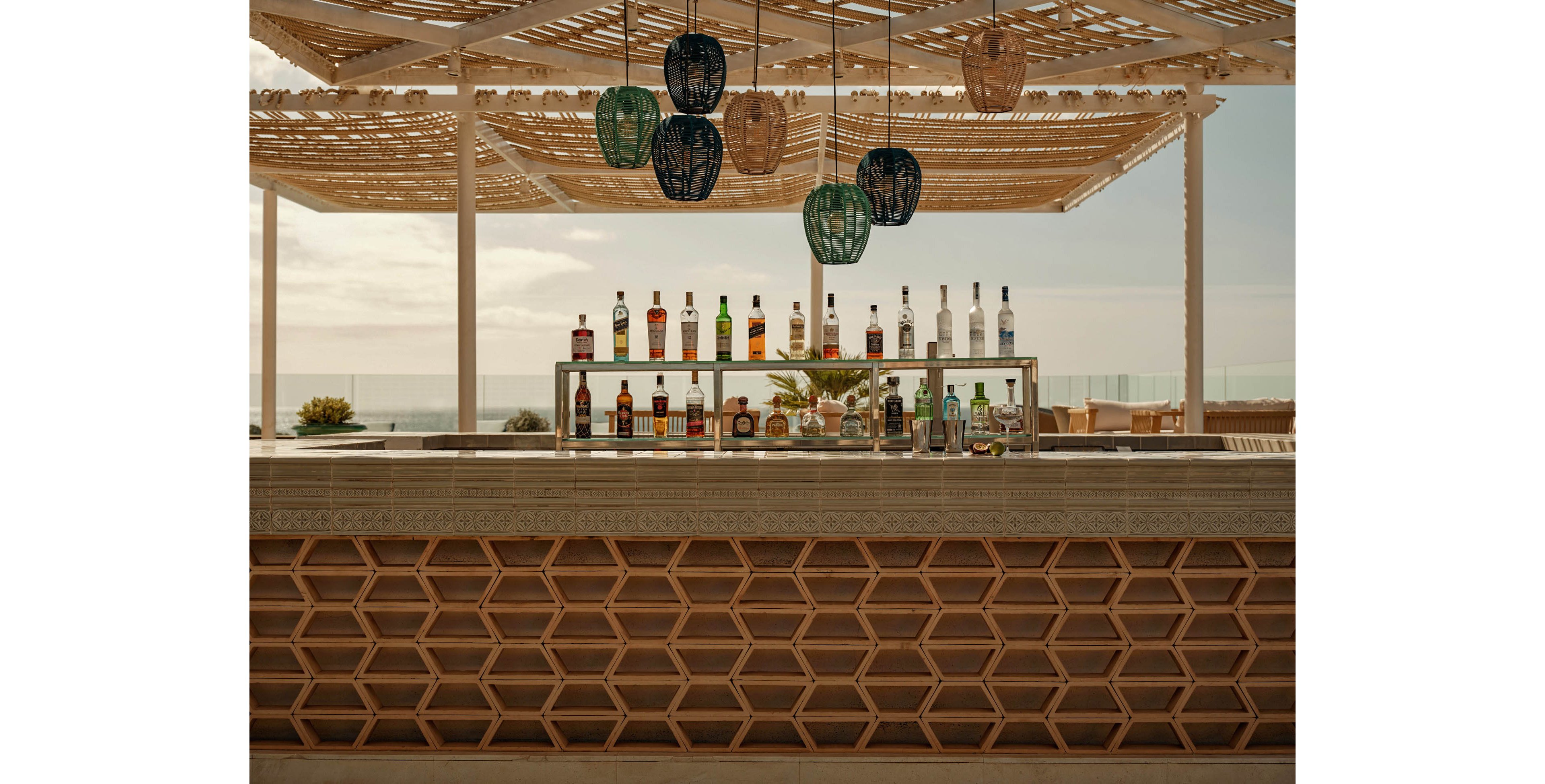 An outdoor bar counter displays a neat row of liquor and spirit bottles on a double-tiered shelf. The bar features a geometric wooden front and a smooth stone countertop. Above, woven pergola slats and hanging lanterns create patterned shade in a coastal setting. The background shows lounge seating and a hazy view of the sea under a soft sky.