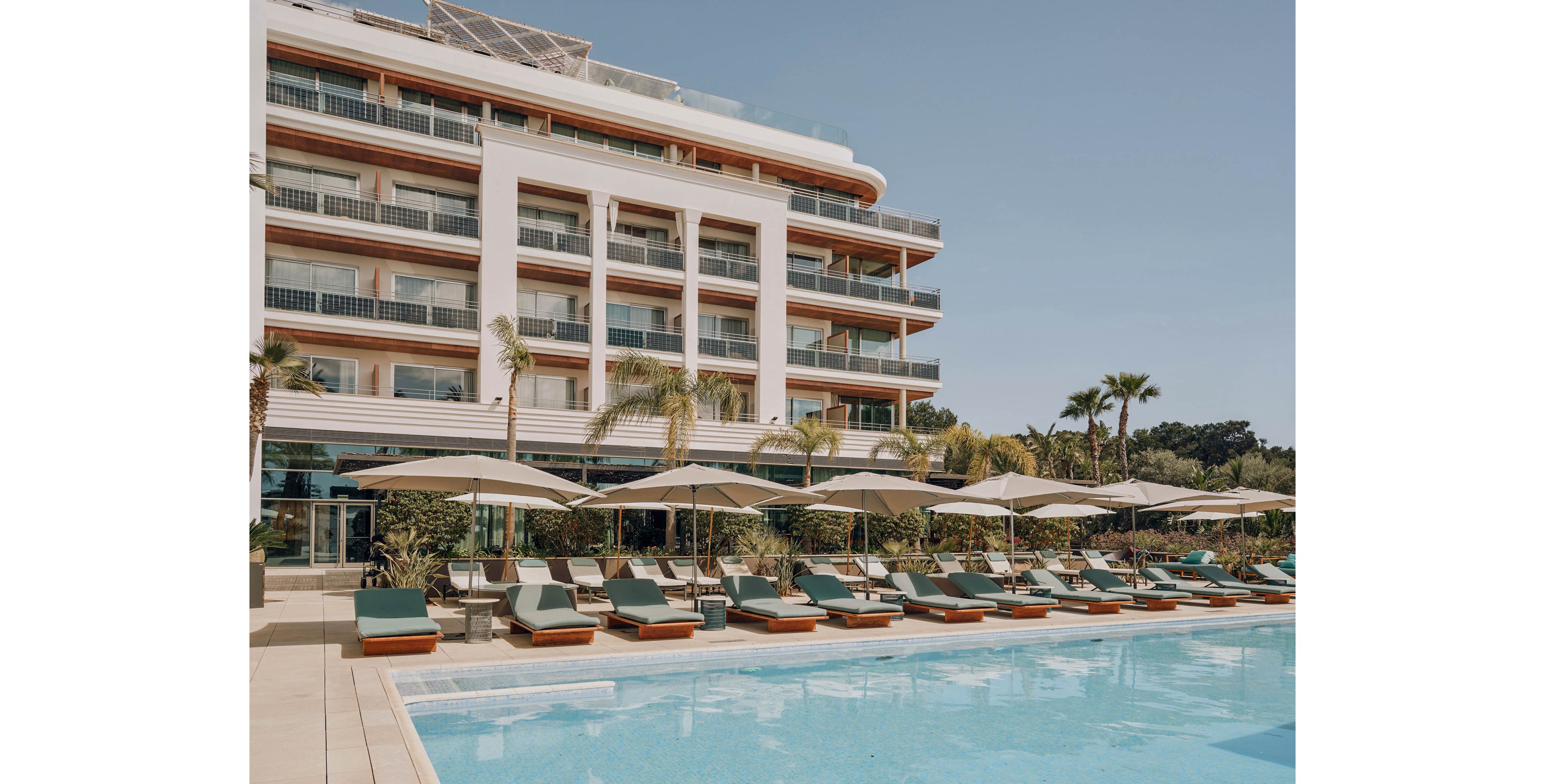 A contemporary multi-story resort hotel overlooks a large outdoor swimming pool. Numerous cushioned sun loungers with umbrellas line the pool deck, creating an inviting relaxation area. Palm trees and lush greenery surround the terrace, enhancing the vacation atmosphere. The clear sky and calm water suggest a warm, sunny day at a luxury destination.