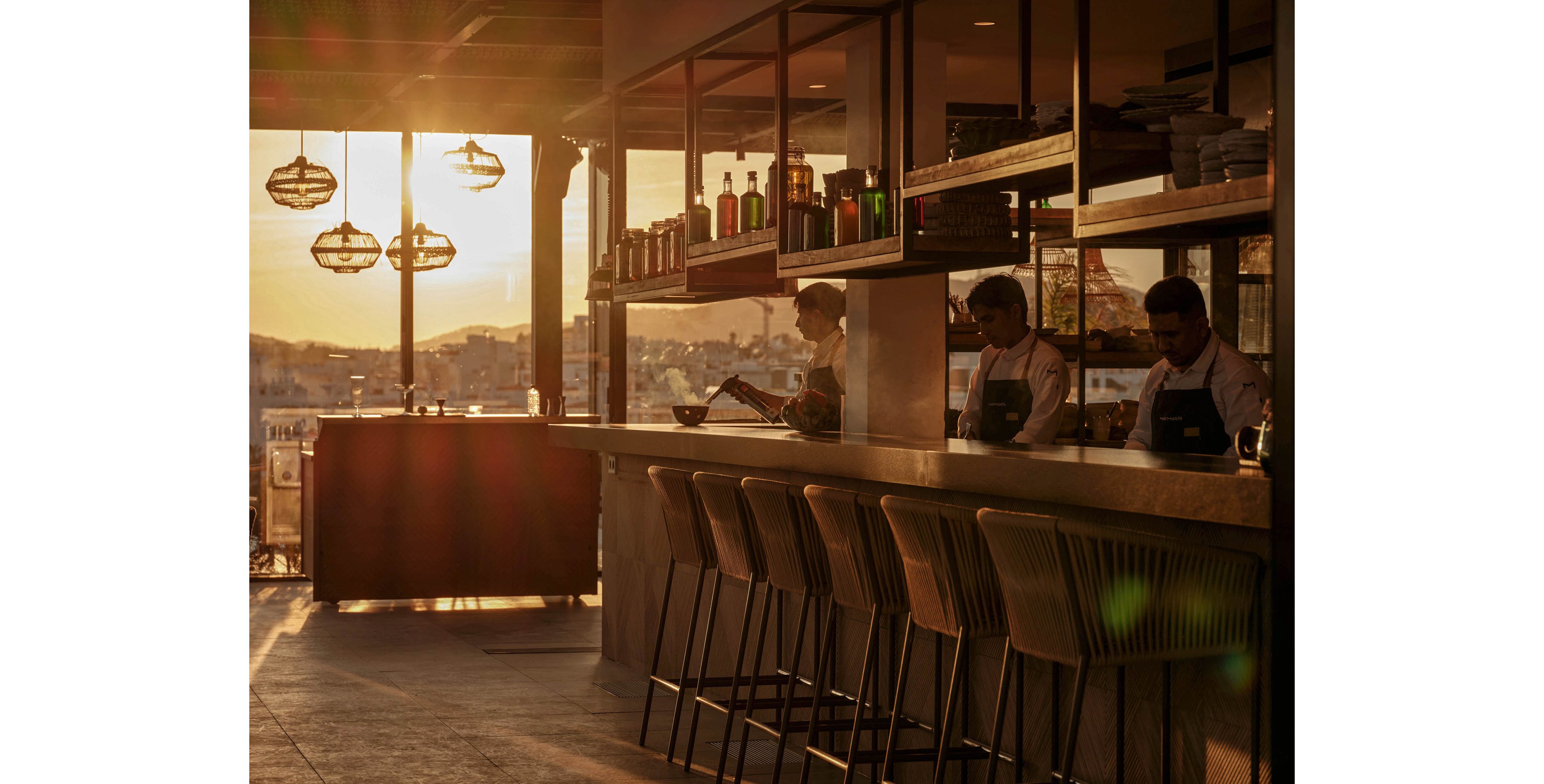 A modern bar interior is bathed in warm sunset light streaming through large windows. Several bartenders in aprons work behind the counter, preparing drinks and organizing bottles on open shelves. Empty bar stools line the front of the counter, inviting guests into the cozy space. Hanging pendant lights and glass bottles are silhouetted against the glowing cityscape outside.