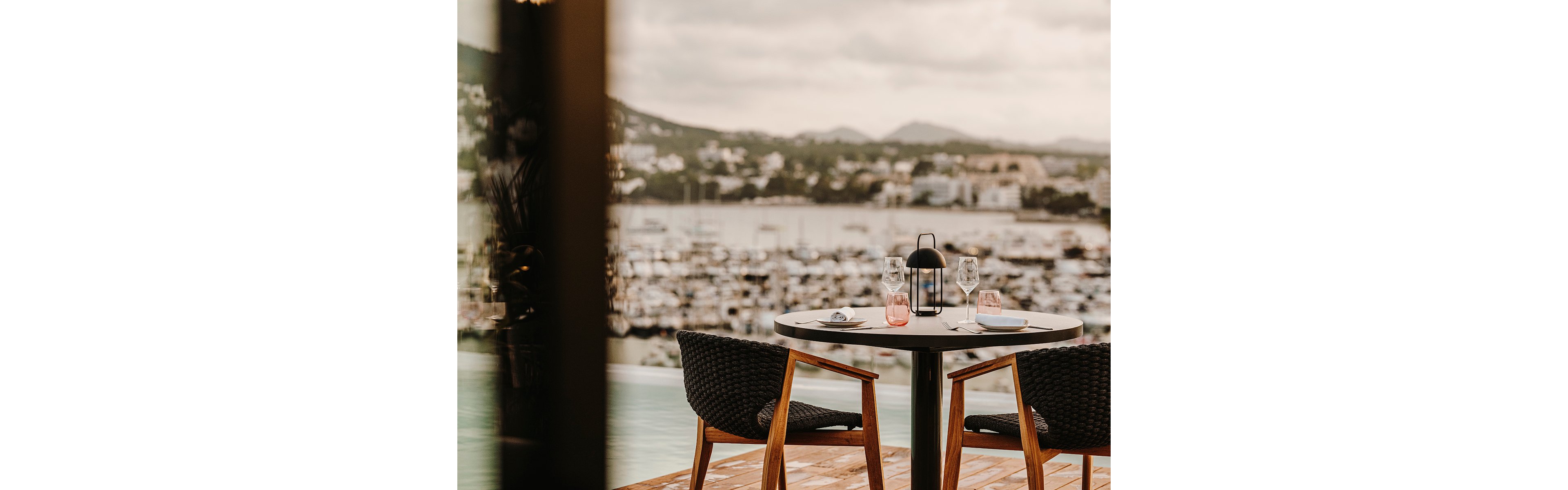 A small round table is set for two on a wooden deck overlooking a marina. Two modern woven chairs face each other with plates, glasses, and pink-tinted tumblers arranged neatly. A black lantern centerpiece adds a cozy, intimate touch to the setting. In the background, numerous boats and a coastal town are softly blurred under an overcast sky.