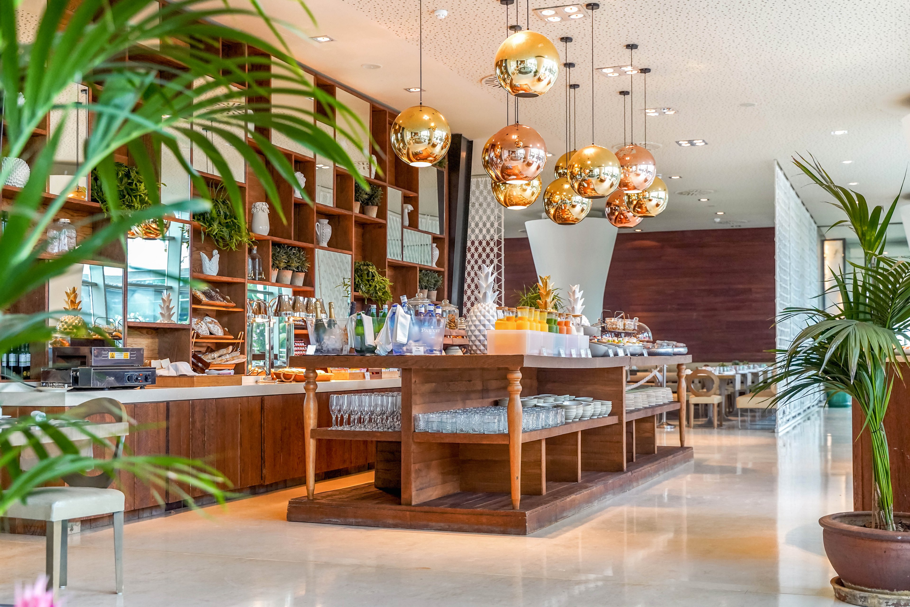 A contemporary café or hotel lounge features a long wooden buffet counter with neatly arranged food and beverage items. Warm metallic pendant lights hang above the counter, reflecting the bright interior. Open wooden shelving with plants and decor lines the back wall, adding a natural touch. Polished floors, indoor greenery, and soft seating create an inviting, upscale atmosphere.