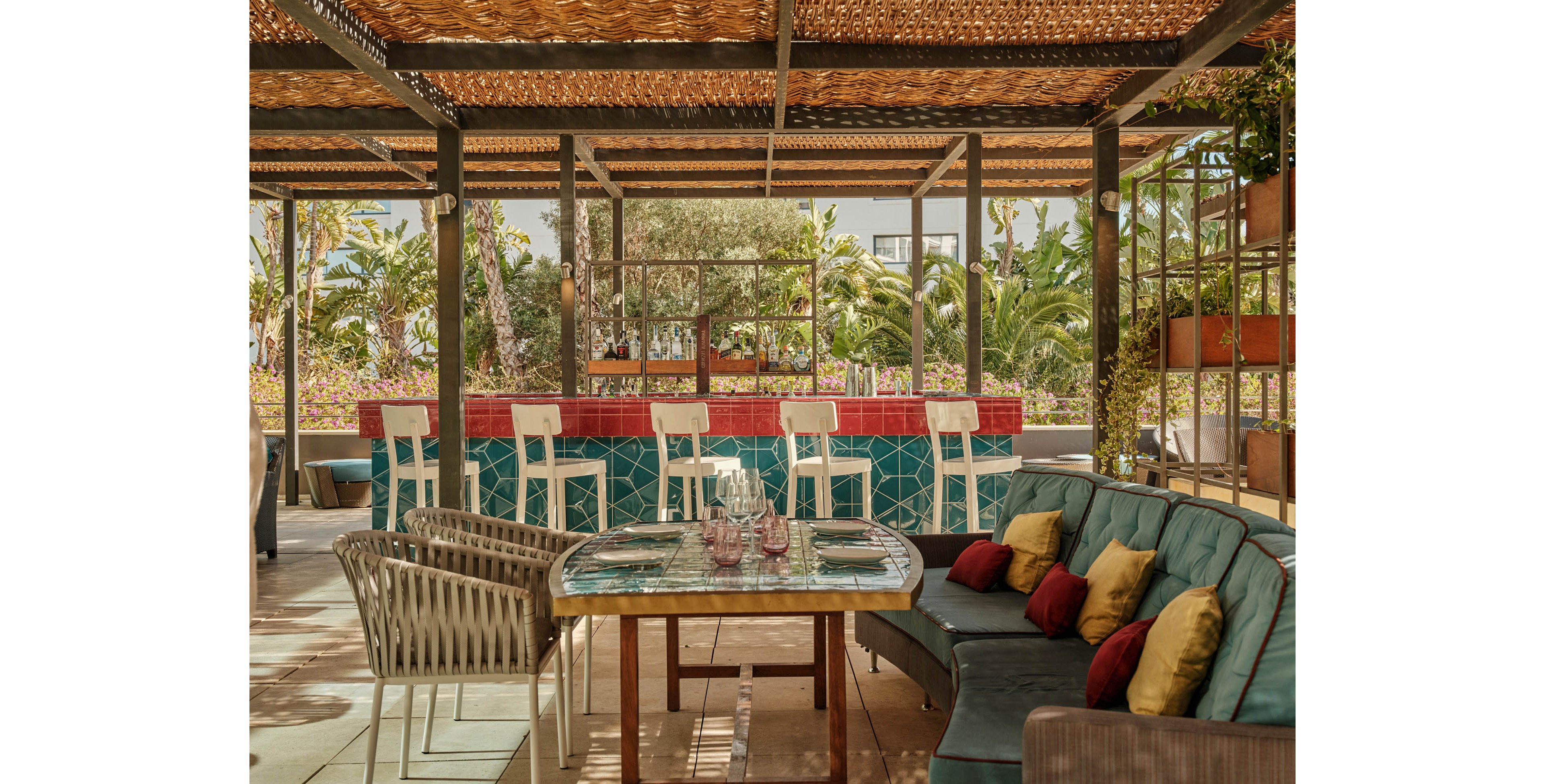 An inviting outdoor bar area features a tiled counter with red and teal geometric patterns and a row of white bar stools. In the foreground, a cozy seating arrangement includes a cushioned sofa with colorful throw pillows and a wicker chair around a mosaic-topped table set with glassware. The space is shaded by a woven pergola roof, creating dappled light on the floor. Lush green plants and palm trees surround the terrace, giving the scene a relaxed, resort-like atmosphere.