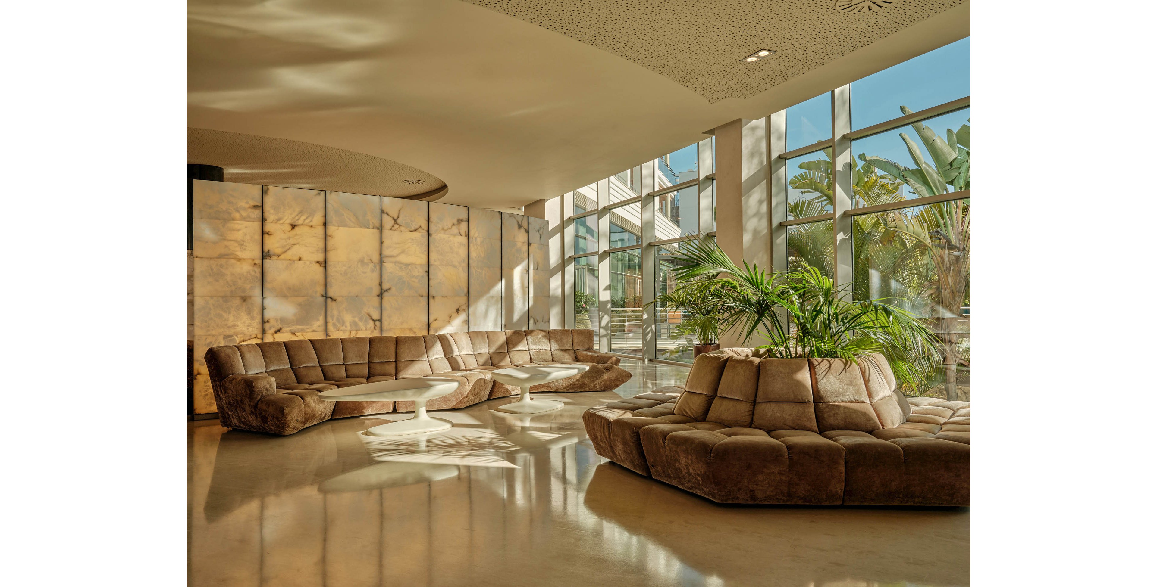 A spacious contemporary lobby features large curved brown sofas arranged around low white tables. Floor-to-ceiling windows flood the room with warm natural light and reveal lush greenery outside. A central planter with tropical plants adds a touch of nature to the sleek interior. The polished floor and backlit stone wall panels create a luxurious, tranquil atmosphere.