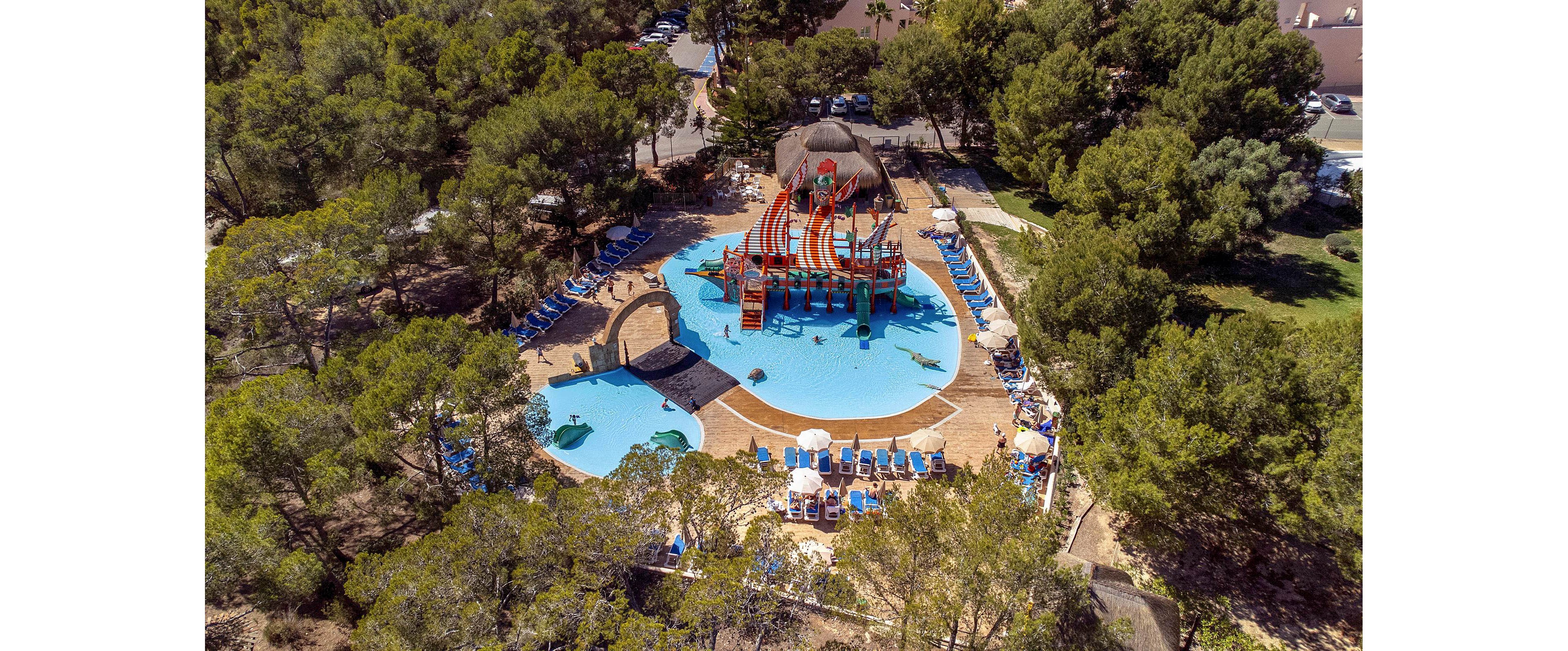 An aerial view shows a colorful outdoor waterpark surrounded by dense green trees. The central structure features multiple bright slides and a shallow splash pool area. Blue lounge chairs and umbrellas are arranged neatly around the pool deck, suggesting a family-friendly resort setting. The scene is sunlit, creating a vibrant and playful summer atmosphere.