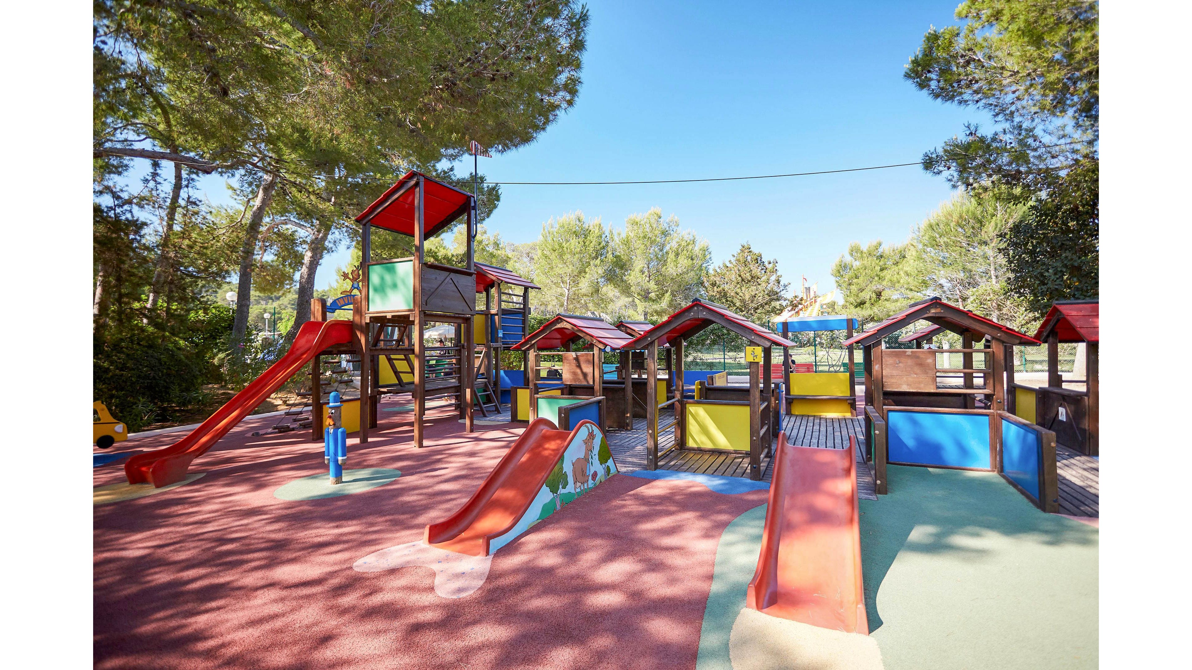 A bright, colorful playground features multiple slides, climbing structures, and small playhouses. The play area is set outdoors, surrounded by tall green trees under a clear sky. Soft rubber flooring in red and green tones covers the ground for safety. The scene is empty of people, highlighting the equipment and open, sunny atmosphere.