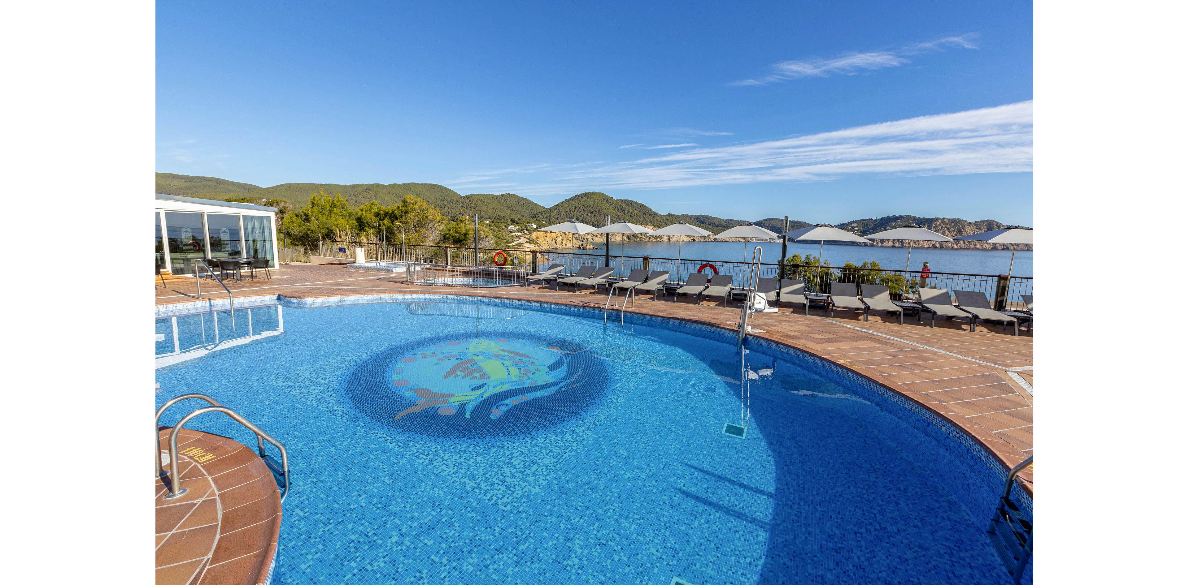 An outdoor resort swimming pool with clear blue water sits on a tiled terrace overlooking a calm coastal bay. Sun loungers and white parasols line the poolside, facing the sea and distant green hills. A decorative circular design is visible on the pool floor, and metal ladders provide access to the water. The sky is bright and clear, creating a relaxed vacation atmosphere.