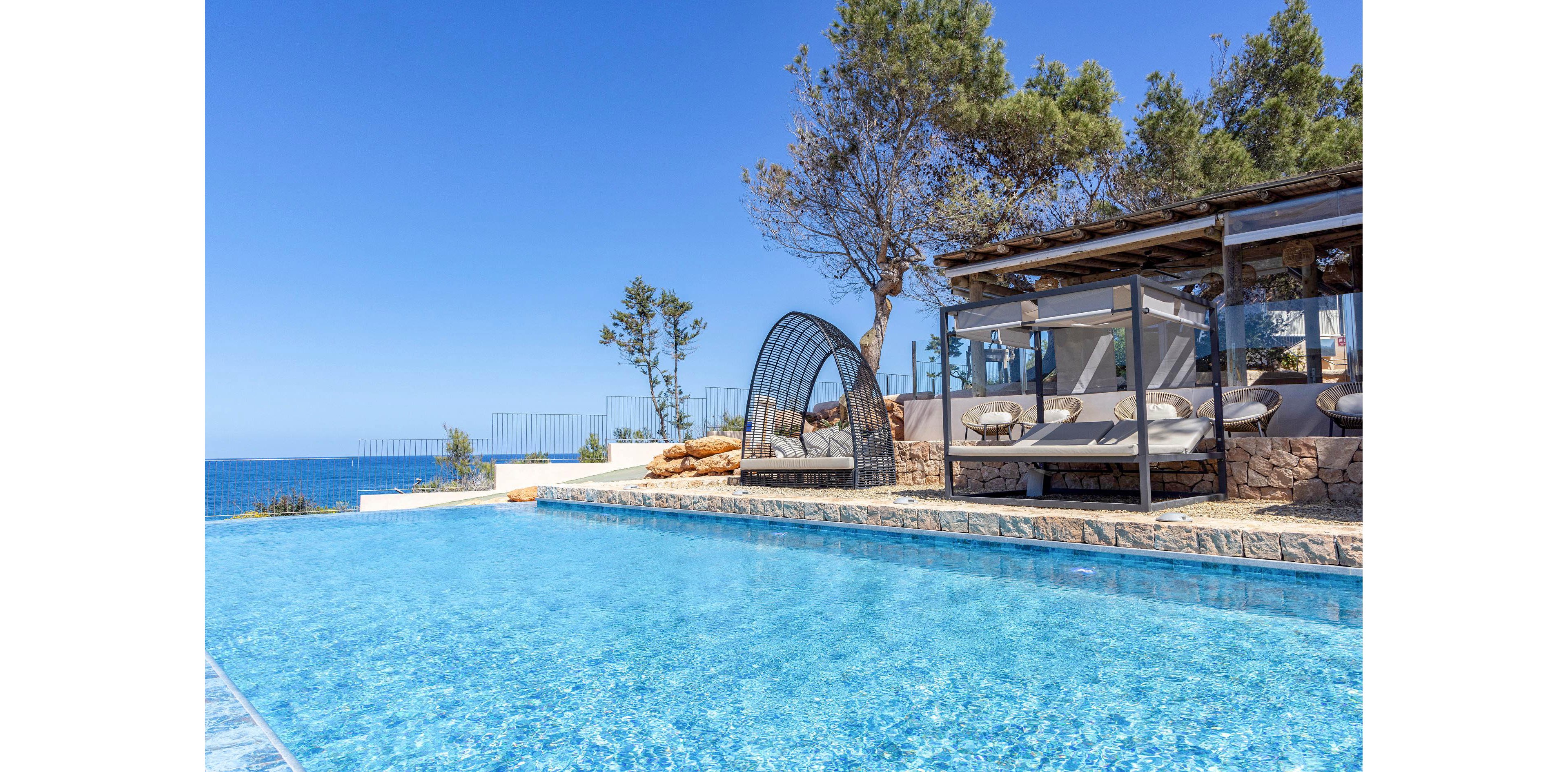 A luxurious infinity pool stretches toward a distant blue sea under a clear sky. Modern sunbeds and a shaded lounge area sit on a stone terrace beside the water. Trees and natural rock elements frame the scene, blending architecture with the coastal environment. The bright sunlight and vivid blue tones create a serene, resort-like atmosphere ideal for relaxation.