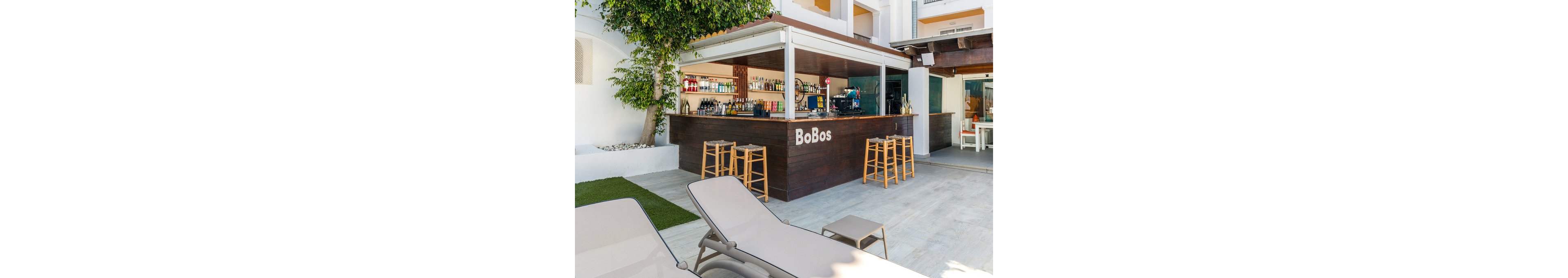 An open-air bar named BoBos features a dark wooden counter and several tall wooden stools. The bar is stocked with various bottles and glassware neatly arranged on shelves. In the foreground, two reclining lounge chairs face the bar, suggesting a relaxed poolside setting. A large tree and modern white building facade frame the scene, creating a bright, summery atmosphere.