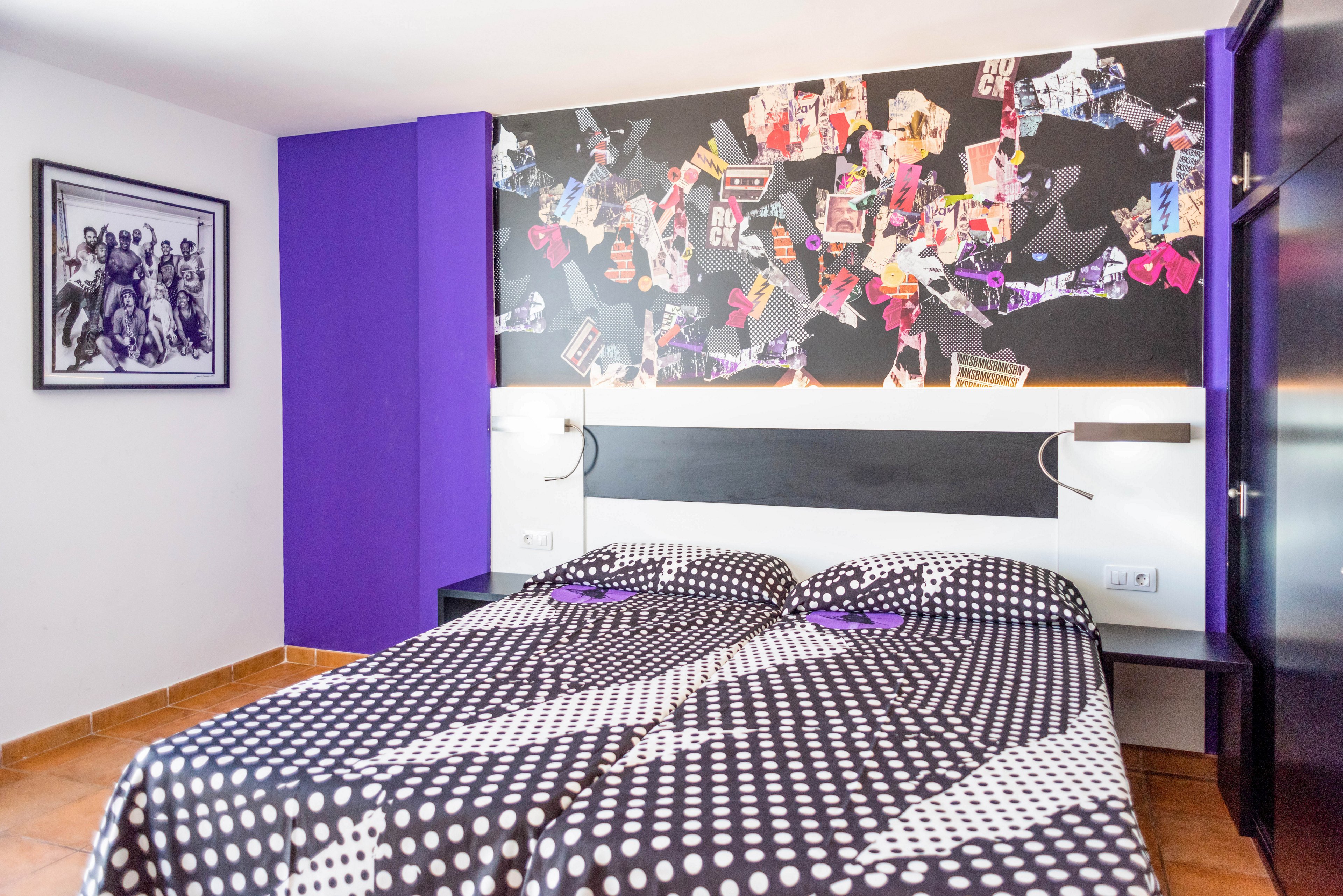 A bright contemporary bedroom features two twin beds with black and white polka dot bedding. The room is accented by a bold purple wall and a vibrant collage-style mural above the headboard. A framed black-and-white artwork hangs on the side wall, adding an artistic touch. Warm tiled flooring and simple furnishings create a clean, modern atmosphere.