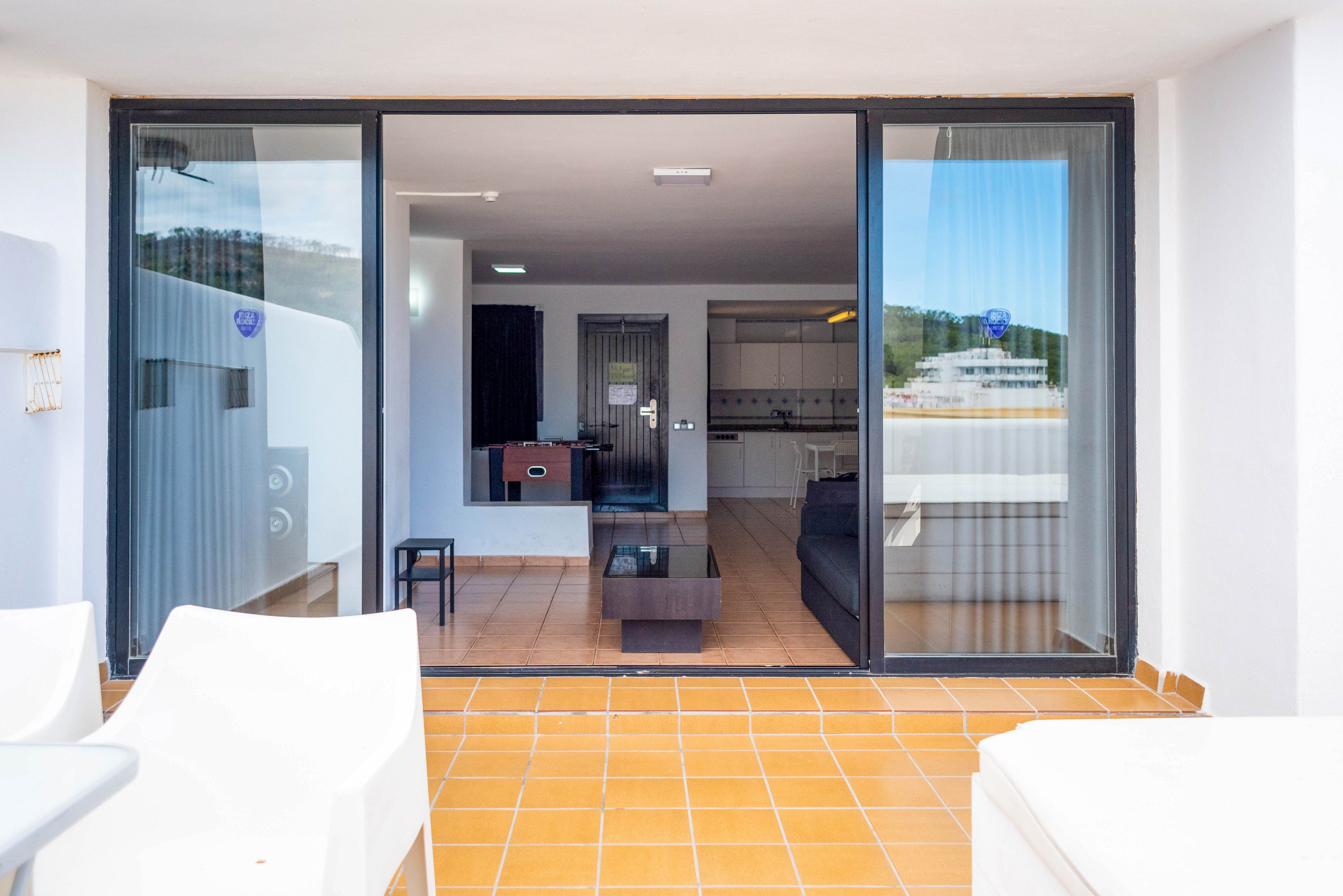 A contemporary apartment living room is viewed from an outdoor terrace through large sliding glass doors. The terrace features tiled flooring and white outdoor furniture, creating a clean and minimal look. Inside, a dark sofa, coffee table, and kitchenette are visible, with neutral colors and simple decor. Reflections in the glass show surrounding buildings and a calm waterfront setting.