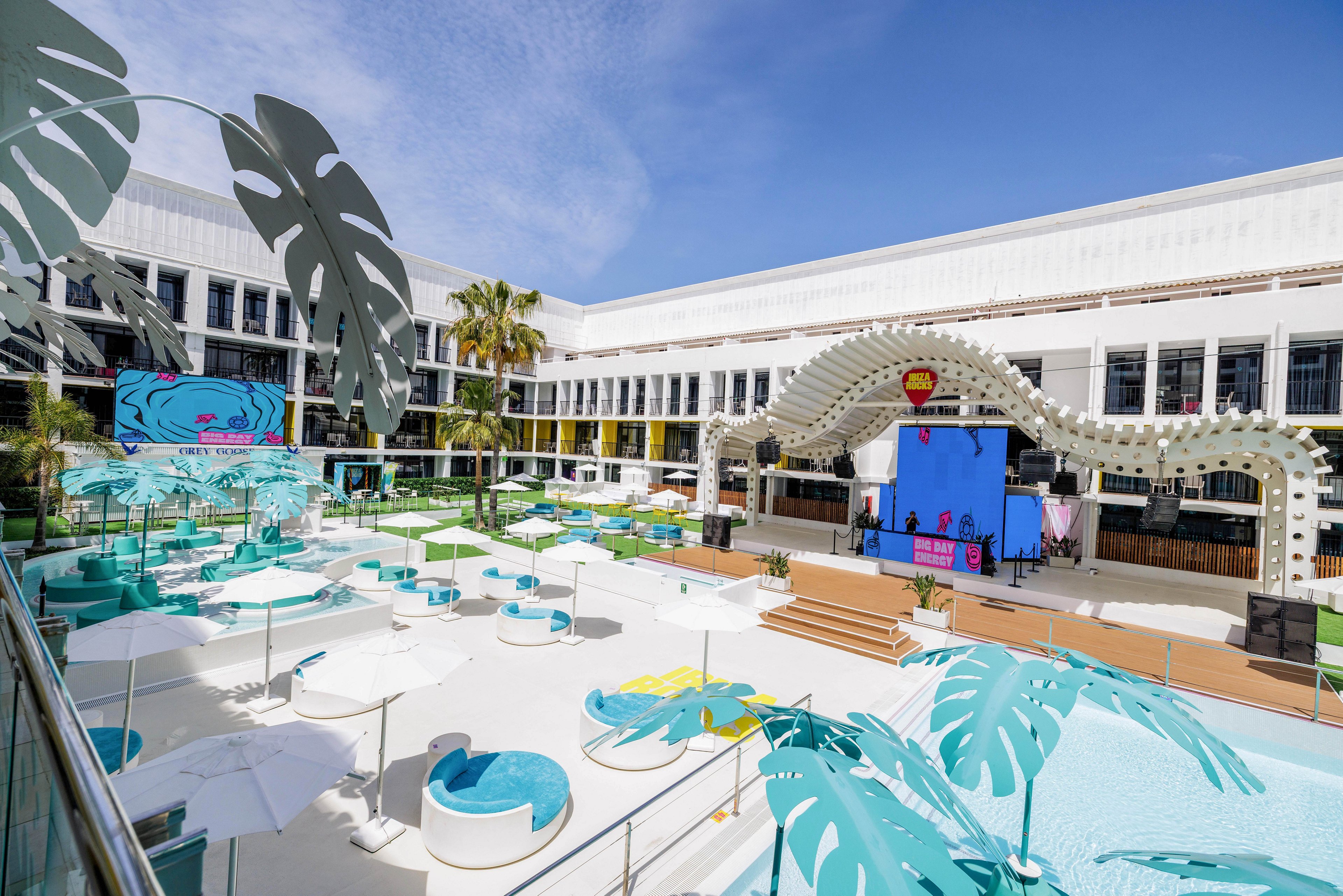 A spacious outdoor pool area is set within a white modern building, featuring turquoise lounge chairs, cabanas, and umbrellas. A large stage with a curved marquee and LED screens is positioned at one end, suggesting an event or entertainment setup. The setting is bright and tropical, with palm trees and oversized decorative leaves adding to the vibrant, summery atmosphere.