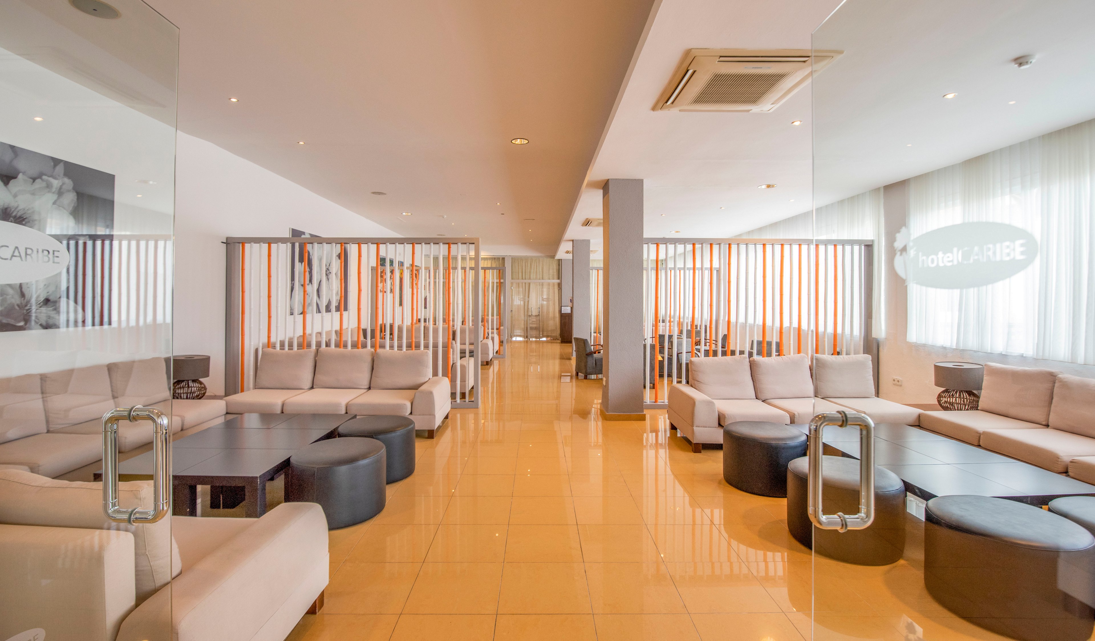 A spacious contemporary hotel lounge features light-colored sofas arranged around low circular tables. Glass partitions with metal handles divide the seating areas, while vertical wooden slats add texture and separation. Large windows with sheer curtains allow natural light to brighten the glossy tiled floor. The space appears calm and inviting, suitable for guests to relax or wait comfortably.