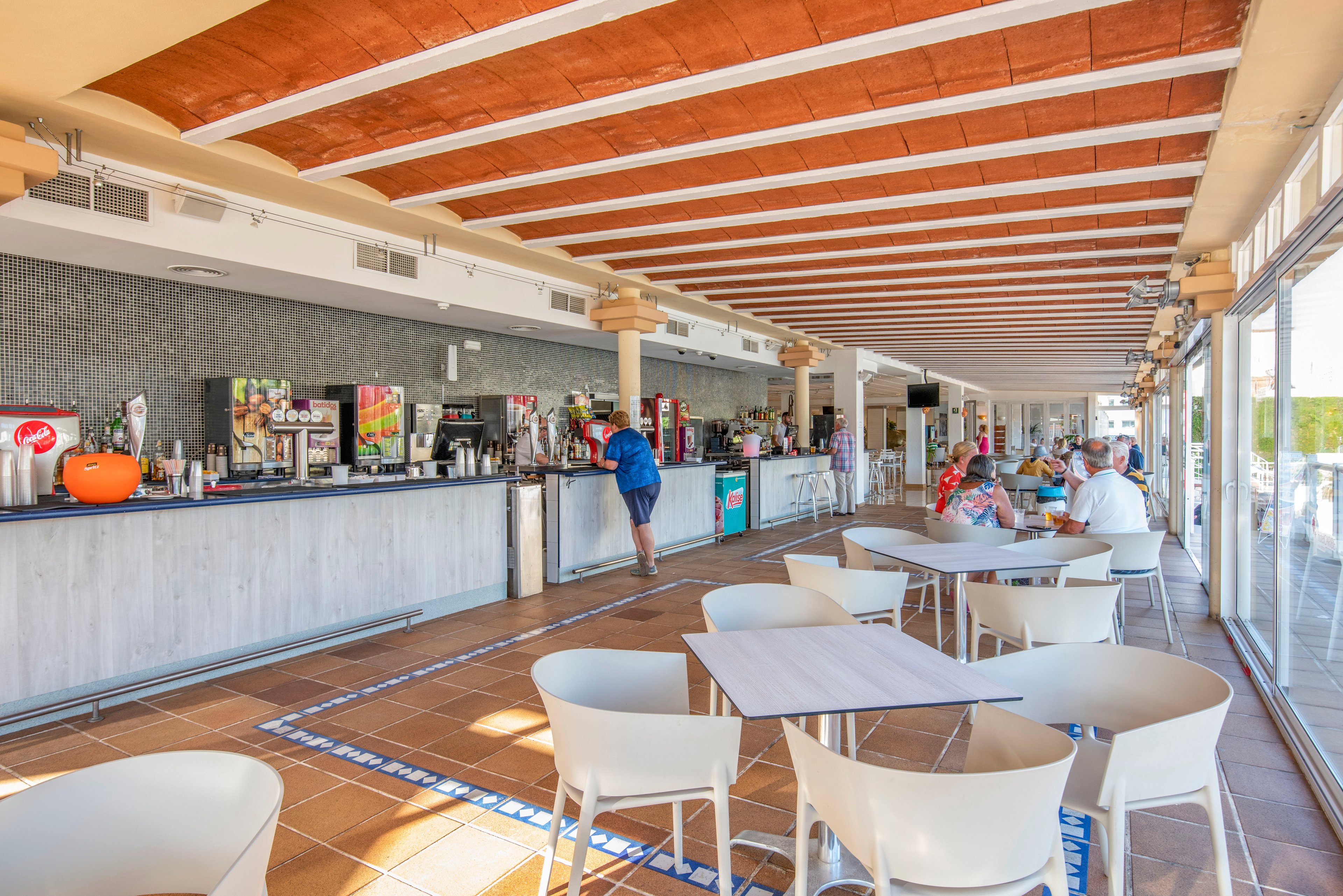 Spacious indoor café bar with large windows shows several people ordering drinks and sitting at tables. White chairs and light wooden tables contrast with the warm tiled floor and exposed beam ceiling. The long counter is lined with beverage dispensers, coffee machines, and colorful drink displays. Sunlight floods the space, creating a relaxed holiday atmosphere suitable for casual dining and refreshments.