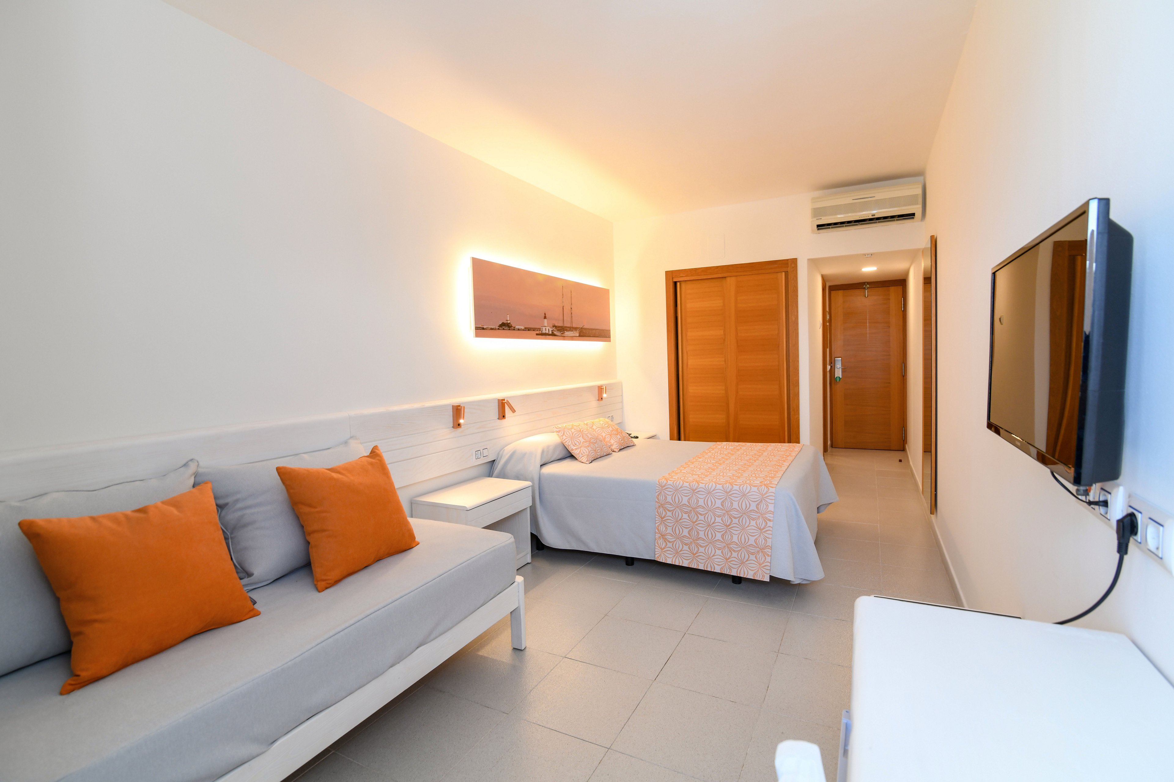 A bright, modern hotel room features a neatly made bed and a light gray sofa with orange accent pillows. Warm recessed lighting and a panoramic wall art piece create a cozy atmosphere. A flat-screen television is mounted on the wall opposite the bed, and wooden doors lead to adjoining areas. The room has clean white walls, tiled flooring, and a simple, minimalist design.
