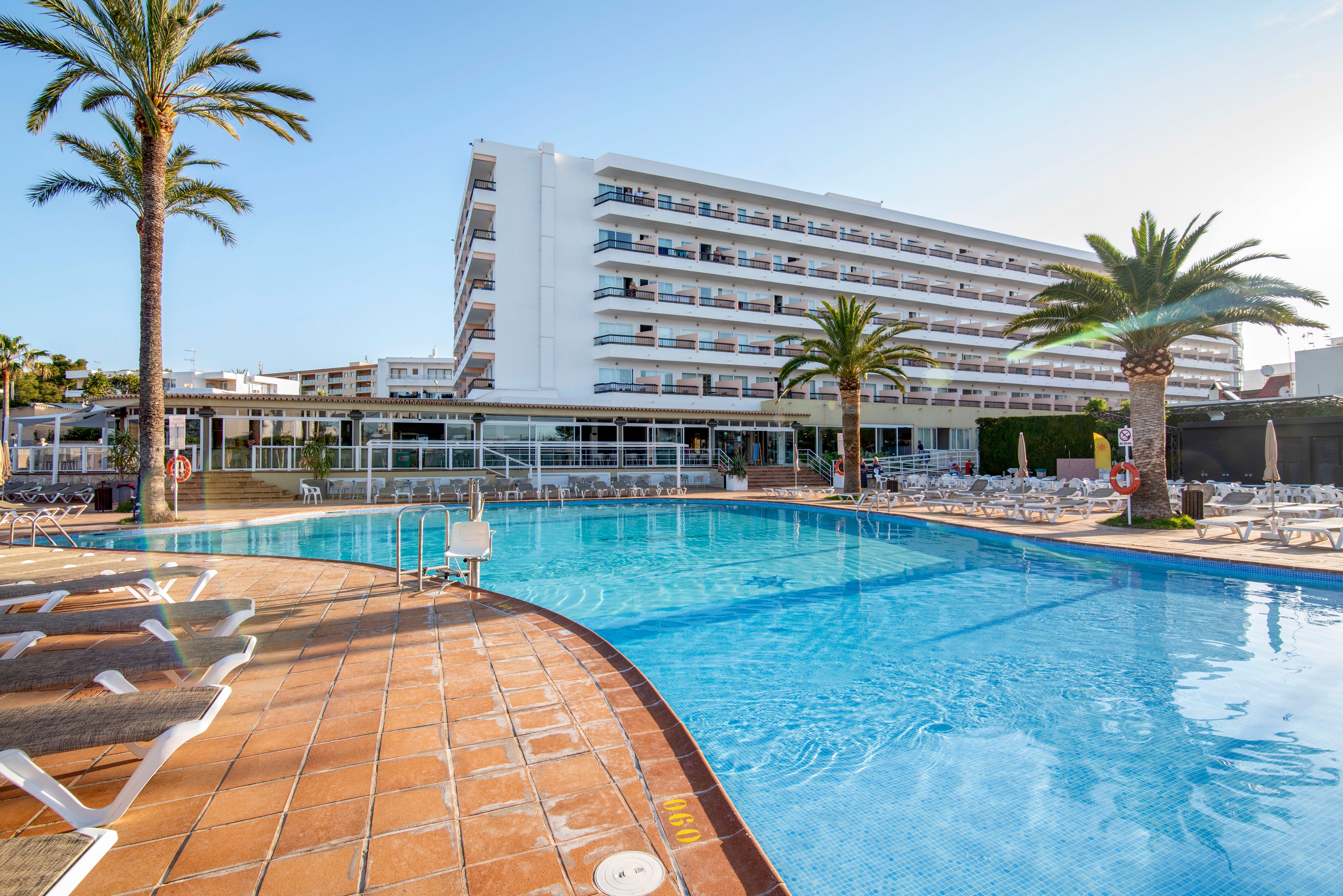 A large multi-story resort hotel overlooks a spacious outdoor swimming pool. Sun loungers line the tiled poolside terrace, with palm trees adding a tropical atmosphere. The clear blue water reflects the bright sky, suggesting a warm, sunny day. The scene conveys a relaxed vacation setting ideal for leisure and relaxation.