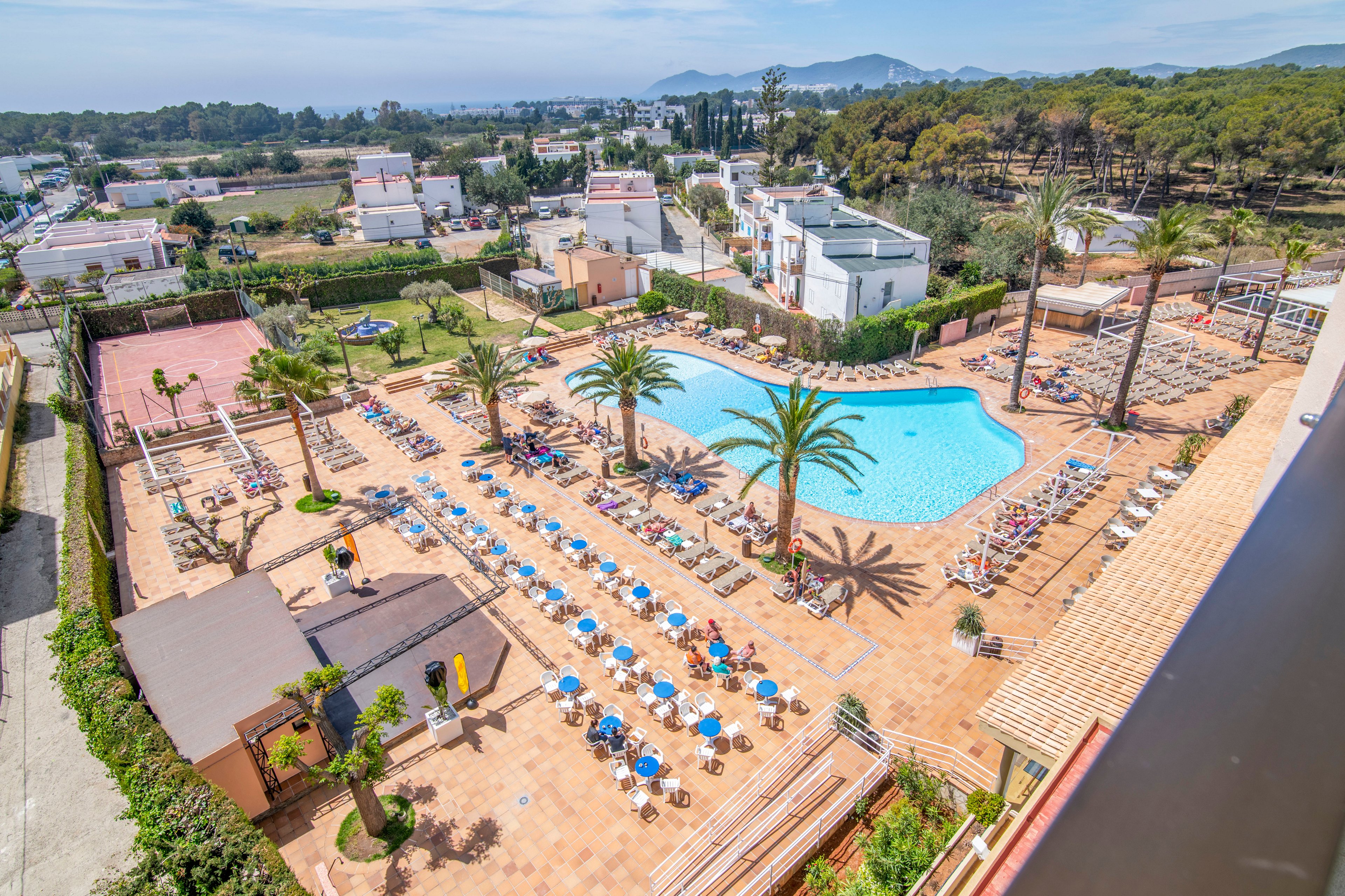 A large outdoor swimming pool is surrounded by rows of sun loungers and blue parasols on a spacious tiled terrace. The resort complex includes nearby white buildings, landscaped palm trees, and a tennis court area. In the background, a forested area and distant mountains frame the scene under a clear blue sky. The setting suggests a warm, sunny holiday destination designed for relaxation and leisure.